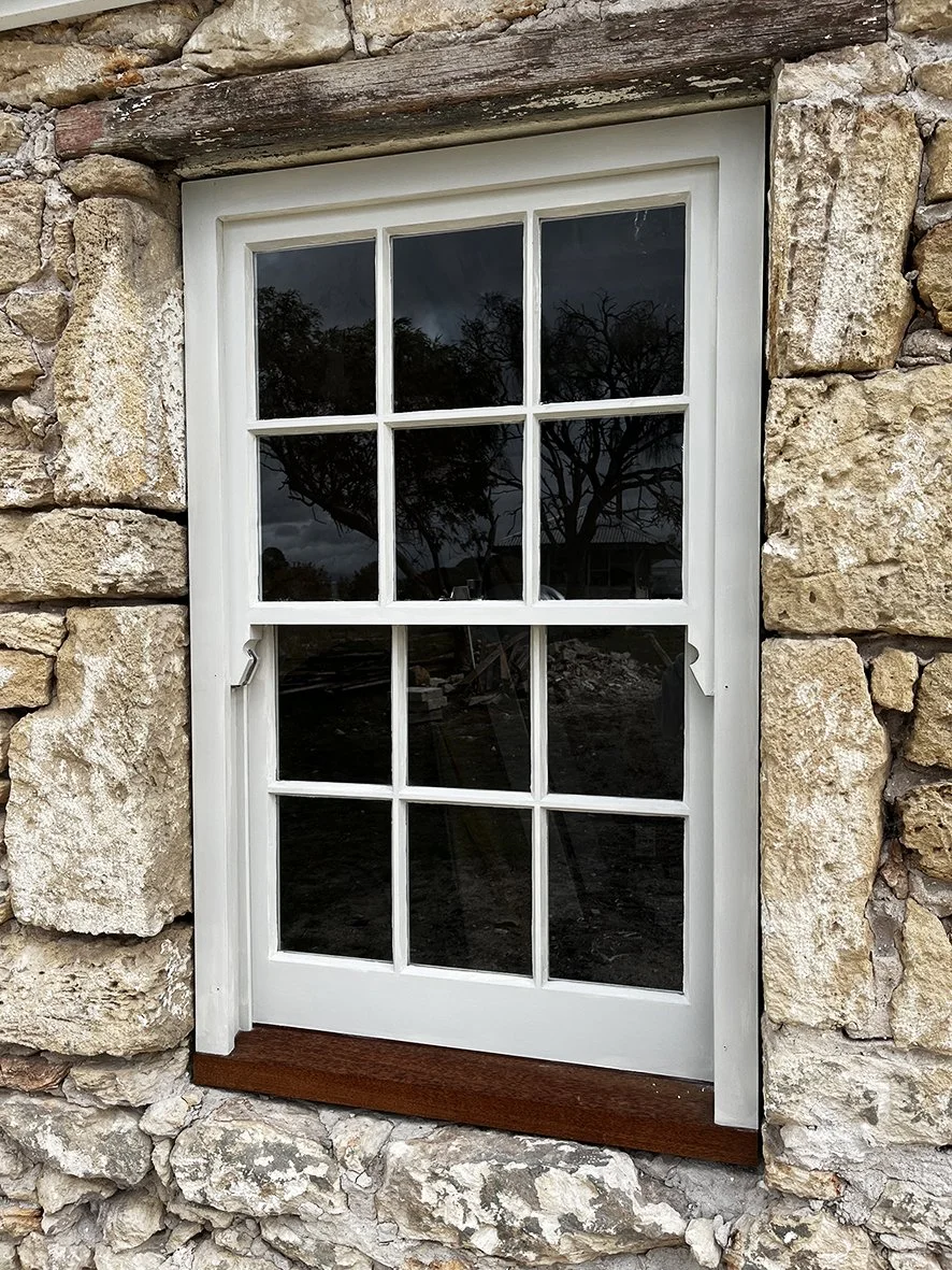 A close-up of a white-framed window with multiple panes, set in a stone wall. The window shows a reflection of trees and a cloudy sky outside.