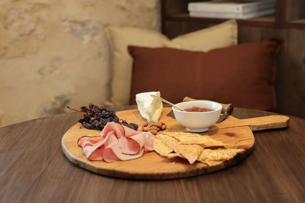 Charcuterie board with dried grapes, sliced prosciutto, cheese wedge, crackers, and a small bowl of sauce or jam.