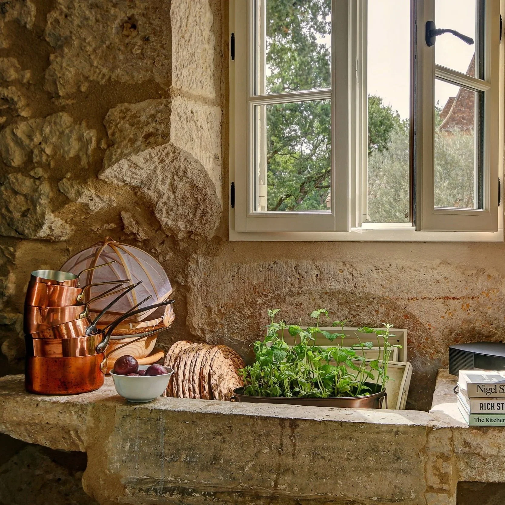 View from inside a rustic kitchen or living space showing a window with a greenery outside, filled with natural light. On the stone countertop, there are copper cookware, a small bowl of purple potatoes, a basket with bread, a plant in a black container, and a stack of books titled "Nigel Slater", "Dick St" and "The Kitchen".