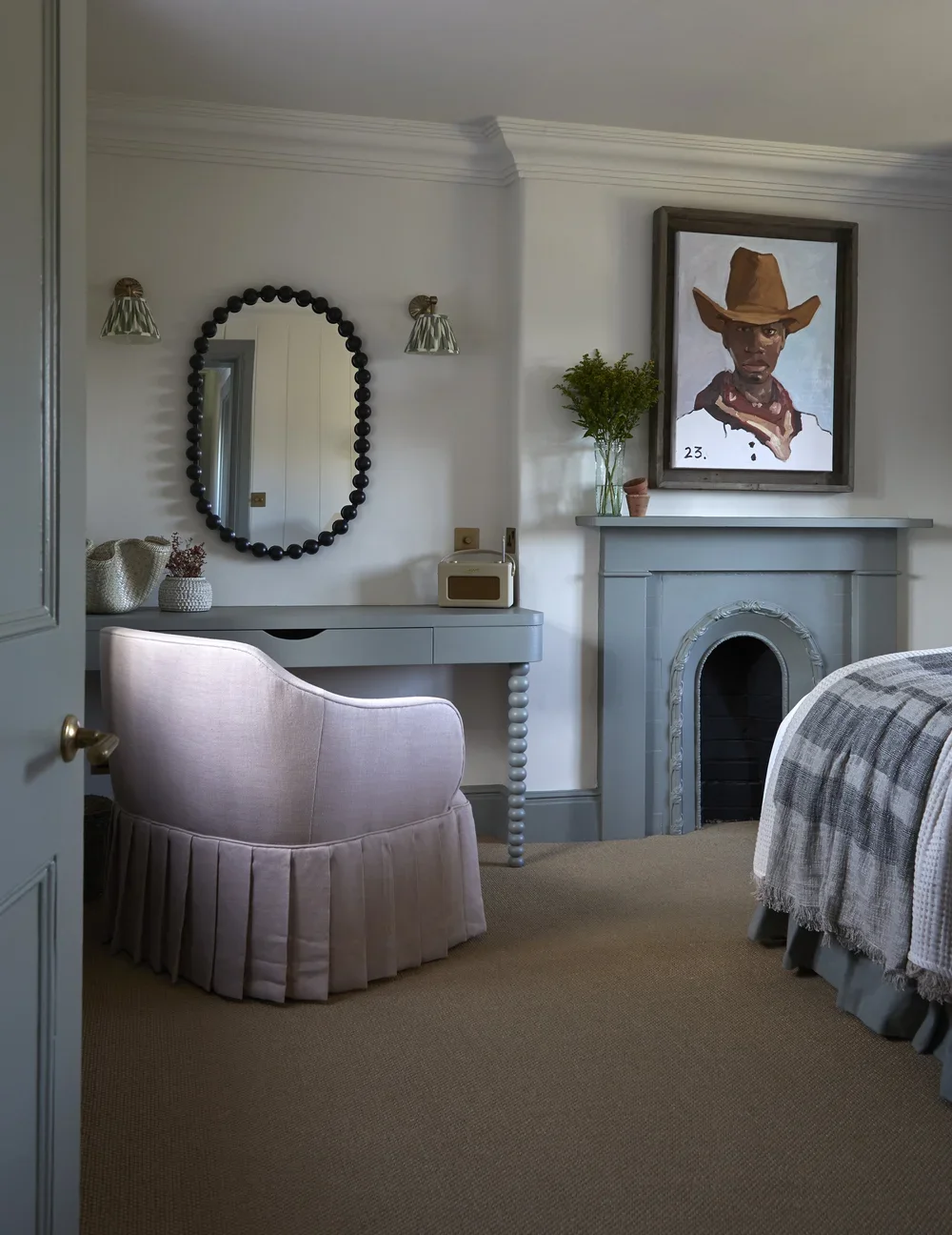 A cozy bedroom corner with a pink upholstered armchair, a light blue desk, and a decorative fireplace. A mirror with a black beaded frame hangs above the desk, flanked by two small wall sconces. On the mantle, there is a vase with greenery, a small potted plant, and a framed portrait of a person wearing a cowboy hat.