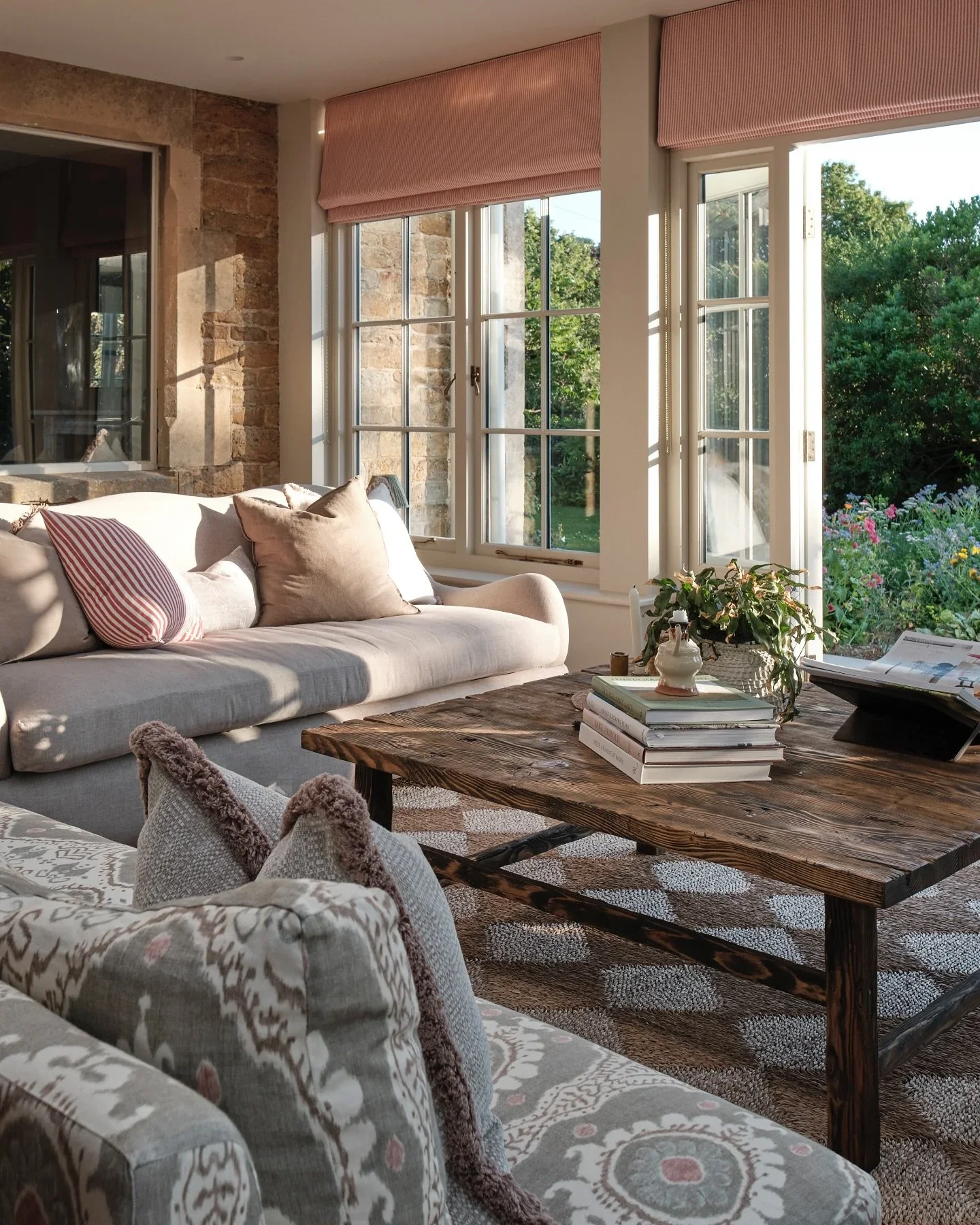 A cozy living room with a beige sofa, decorative pillows, a rustic wooden coffee table, and large windows that let in natural sunlight and offer a view of a garden with green trees and colorful flowers.