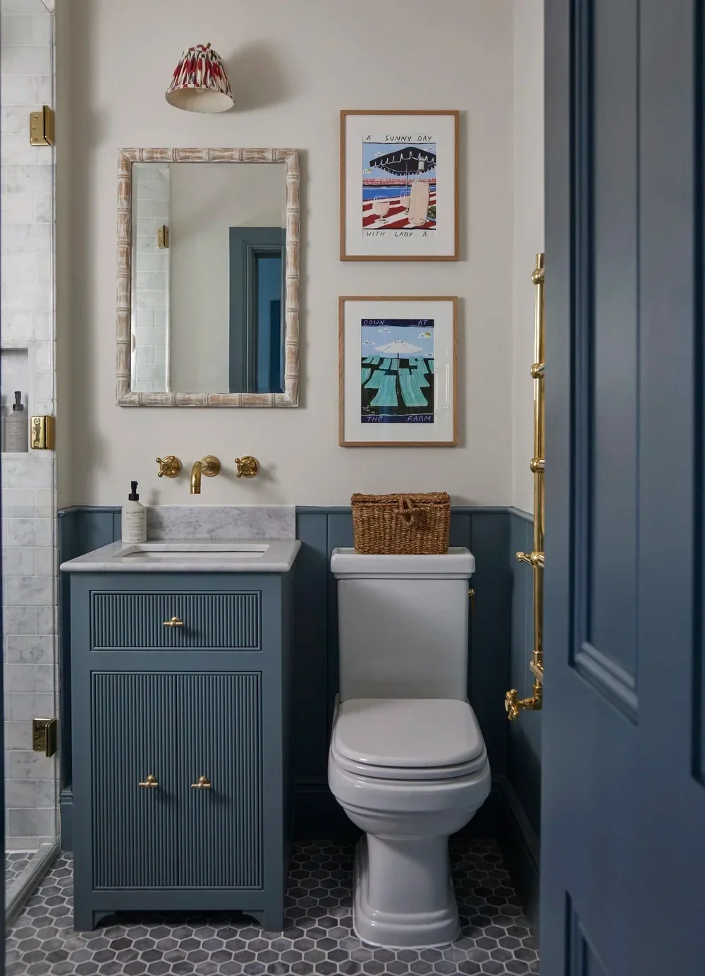 A small bathroom with a blue vanity with a white marble top, gold fixtures, and a white toilet with a wicker basket on top. The wall features two framed art prints and a mirror. The flooring is gray hexagonal tile, and part of a shower with marble tiles and gold hardware is visible.