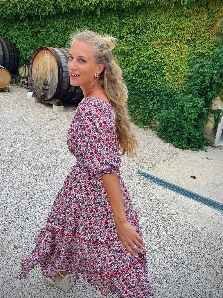 Woman in a long floral dress with curly blonde hair standing outdoors on gravel, with large wine barrels and green ivy in the background.