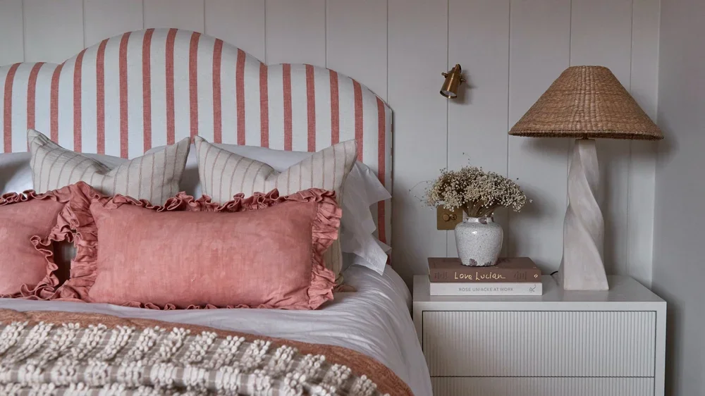 A cozy bedroom corner with a bed featuring a pink and white striped headboard, patterned pillows, and a ruffled pink pillow. A white nightstand holds a vase of baby's breath flowers, a small stack of books, and a lamp with a woven shade. The wall behind is white with paneling, and a brass wall light is mounted above the nightstand.