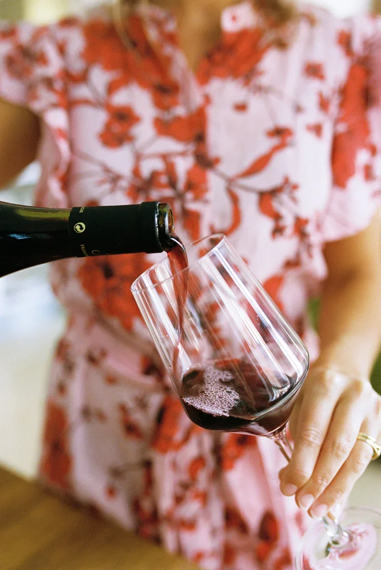 Person in floral dress pouring red wine into a wine glass.