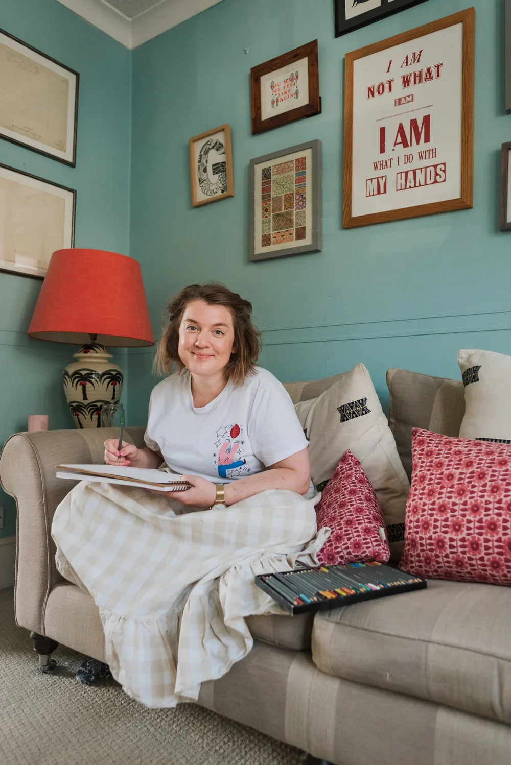 A woman sitting on a beige sofa, smiling, holding a notebook and pen. She is surrounded by colorful pillows and in front of her is a set of colored markers. The living room has teal walls with framed artwork and quotes, a red lampshade on a decorative lamp, and a cozy, stylish ambiance.