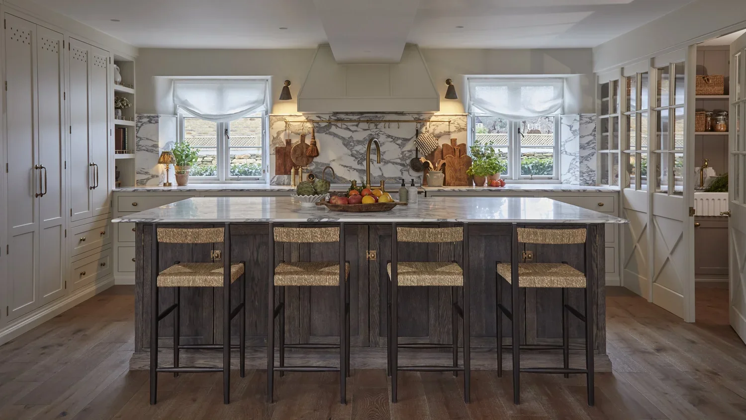 A kitchen with white cabinets, a marble backsplash, a kitchen island with a marble countertop, four bar stools with woven seats, and two windows with white curtains.