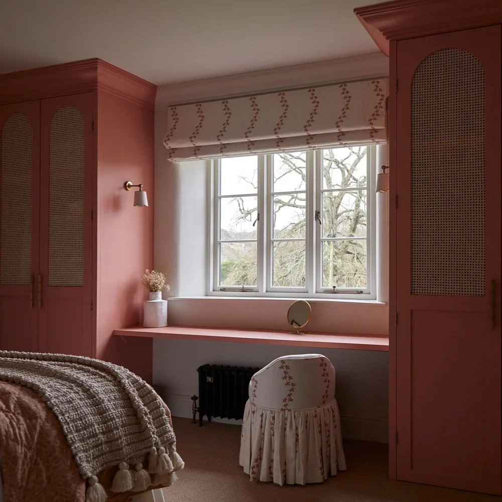 A cozy bedroom corner with pink cabinetry, a window with a floral roman shade, a small vanity with a mirror, and a bed with a textured blanket.