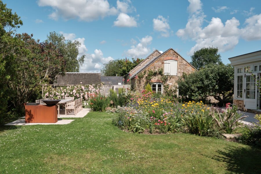 A lush garden with colorful flowers, green bushes, and trees, featuring an outdoor dining area with a table and chairs, and a brick house with a basketball hoop on the wall against a partly cloudy sky.
