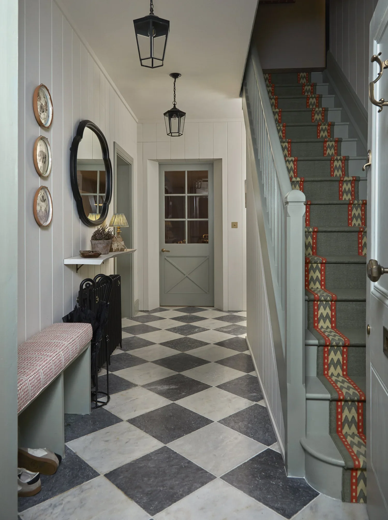 Interior view of a hallway with black and white checkered flooring, white panel walls, and a staircase with decorative carpet runner. Features include a wall mirror, small table with a lamp and potted plant, coat hooks, and three hanging pendant lights.