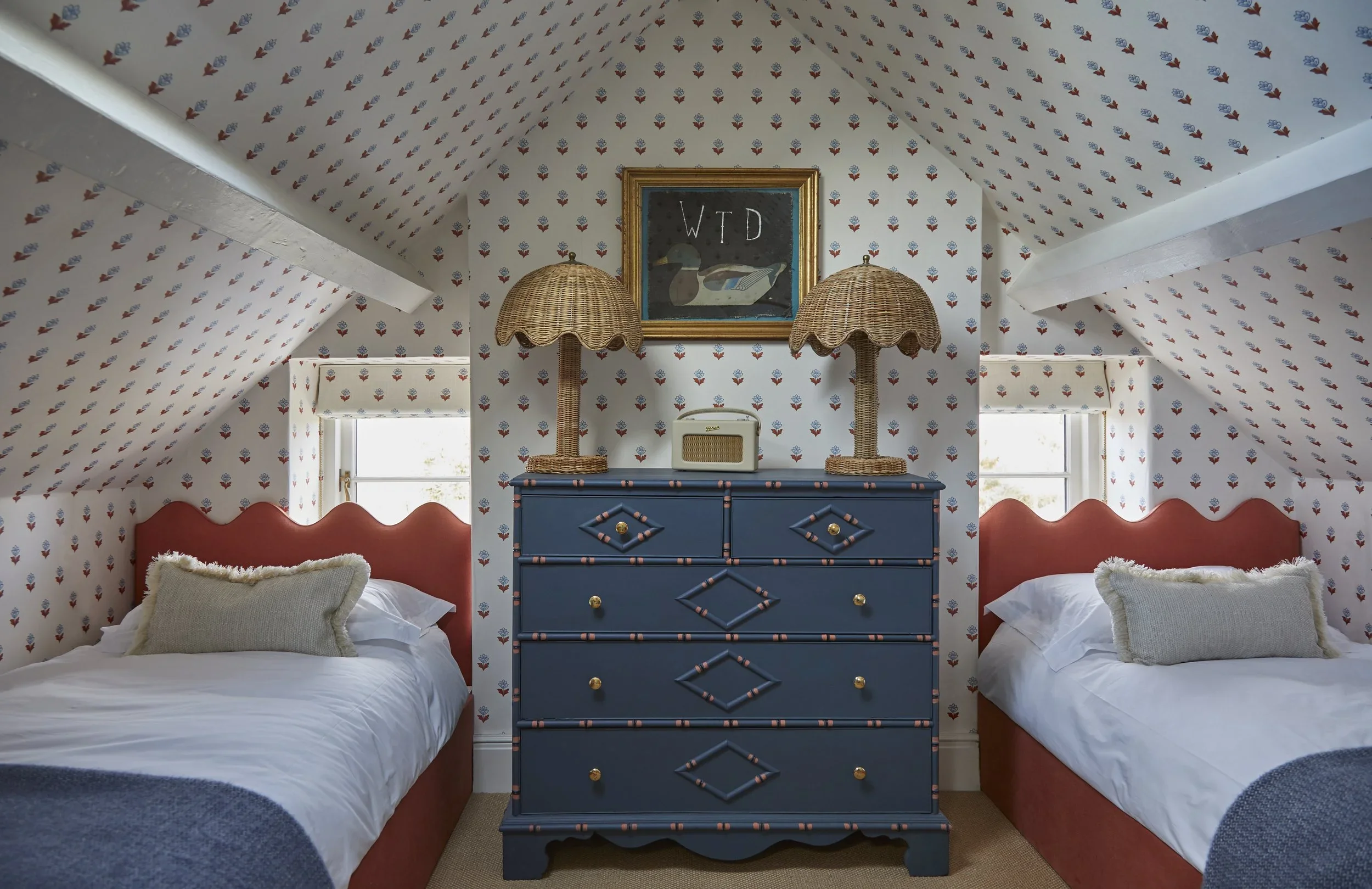 A bedroom with sloped ceiling covered in wallpaper with small red, blue, and white floral patterns. Two beds with red headboards and white bedding are on either side of a blue dresser. The dresser has diamond and striped patterns and gold knobs. Two wicker lamps with scalloped shades sit on the dresser, flanking a framed picture that displays the letters 'WTD' and a duck painting. Small windows with white trim are on either side of the dresser.