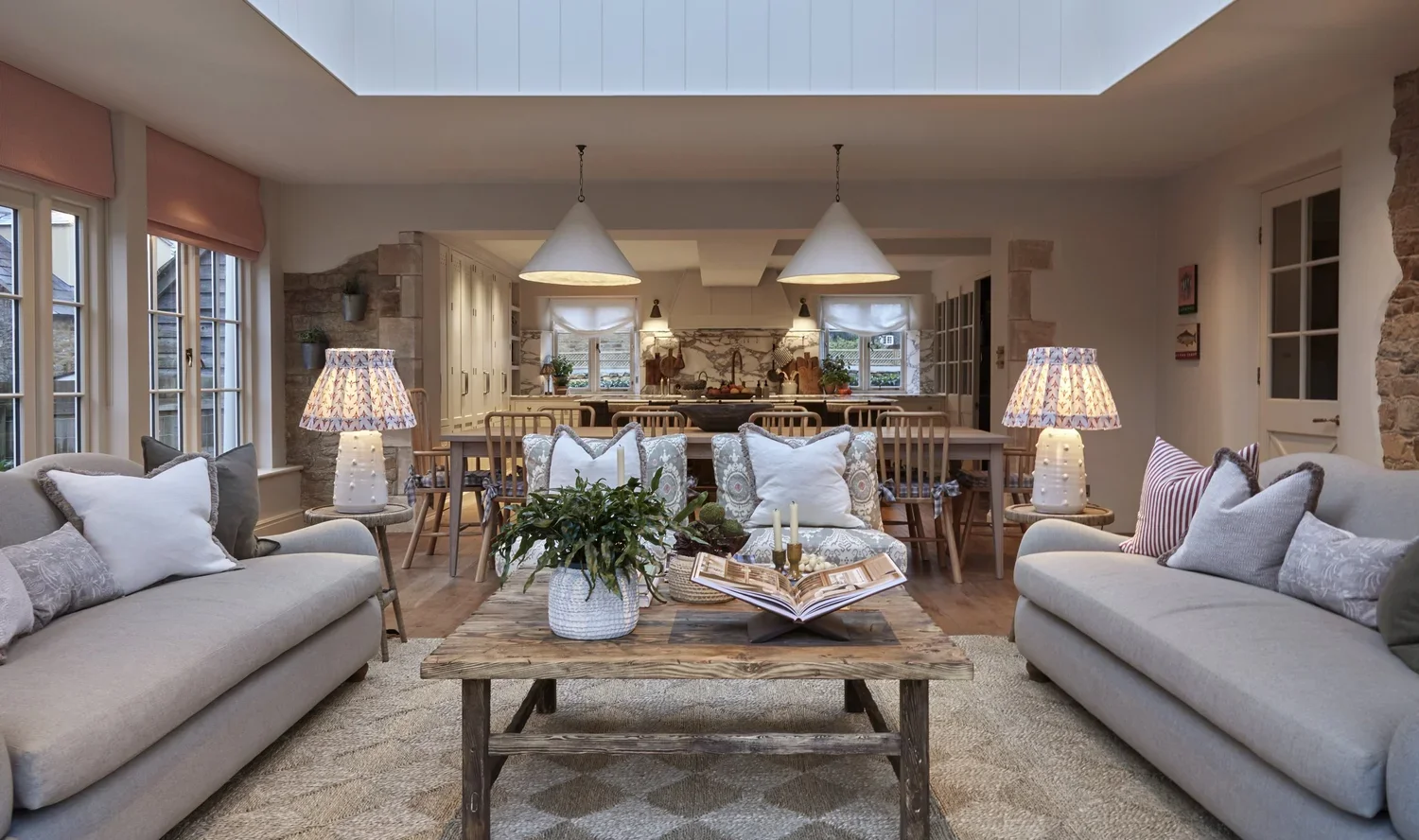 Living room with beige sofas, striped and solid pillows, two lamps with patterned shades on side tables, wooden coffee table with plant and candles, view into dining area with six chairs, pendant lights, and kitchen with windows and stone accents.