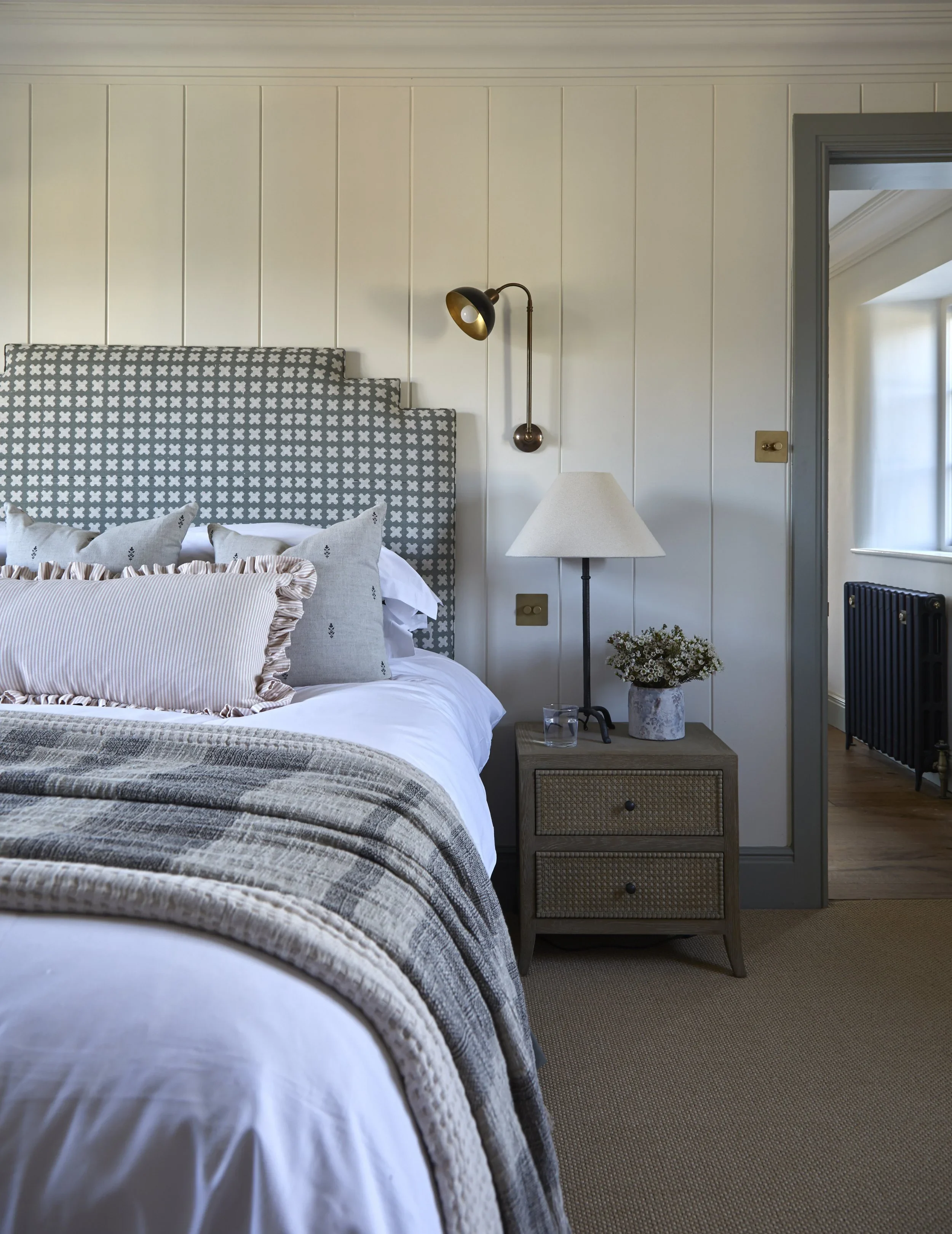 A bedroom with a bed featuring a patterned headboard and decorative pillows, a bedside table with a vase of flowers, a standing lamp, and a wall-mounted reading light