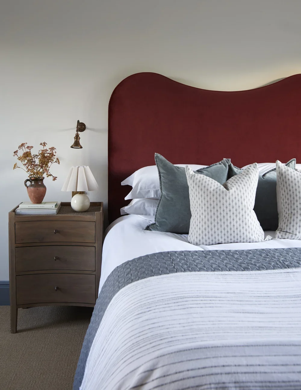 A neatly made bed with a red upholstered headboard, white bedding, and multiple throw pillows in shades of gray and white. A wooden nightstand holds a white lamp, a vase with dried flowers, and a few books. A brass reading light is mounted on the wall above the nightstand.