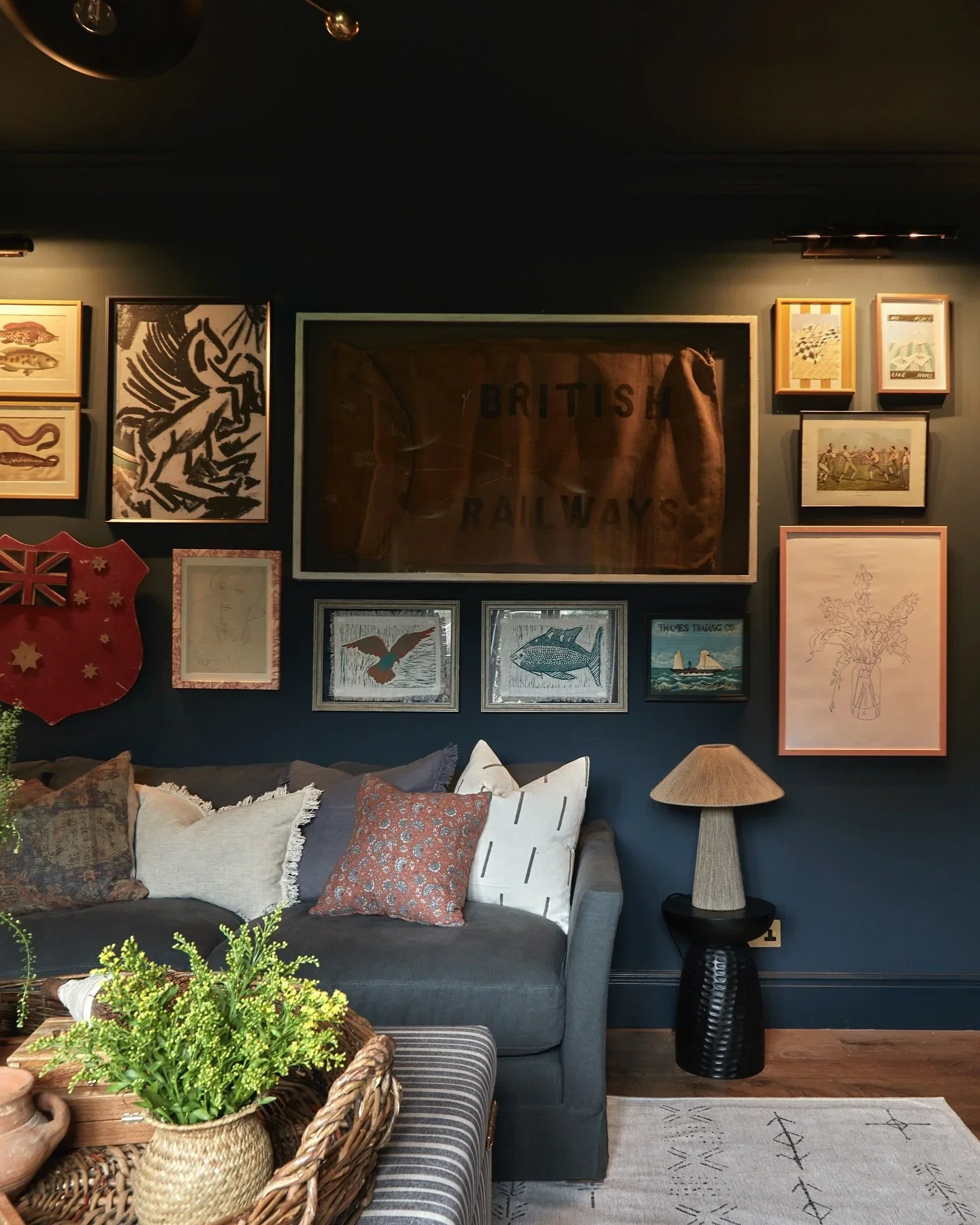 Living room with a gray sofa, various pillows, a side table with a lamp, and a wall decorated with multiple framed art pieces and a framed British Railway sign.