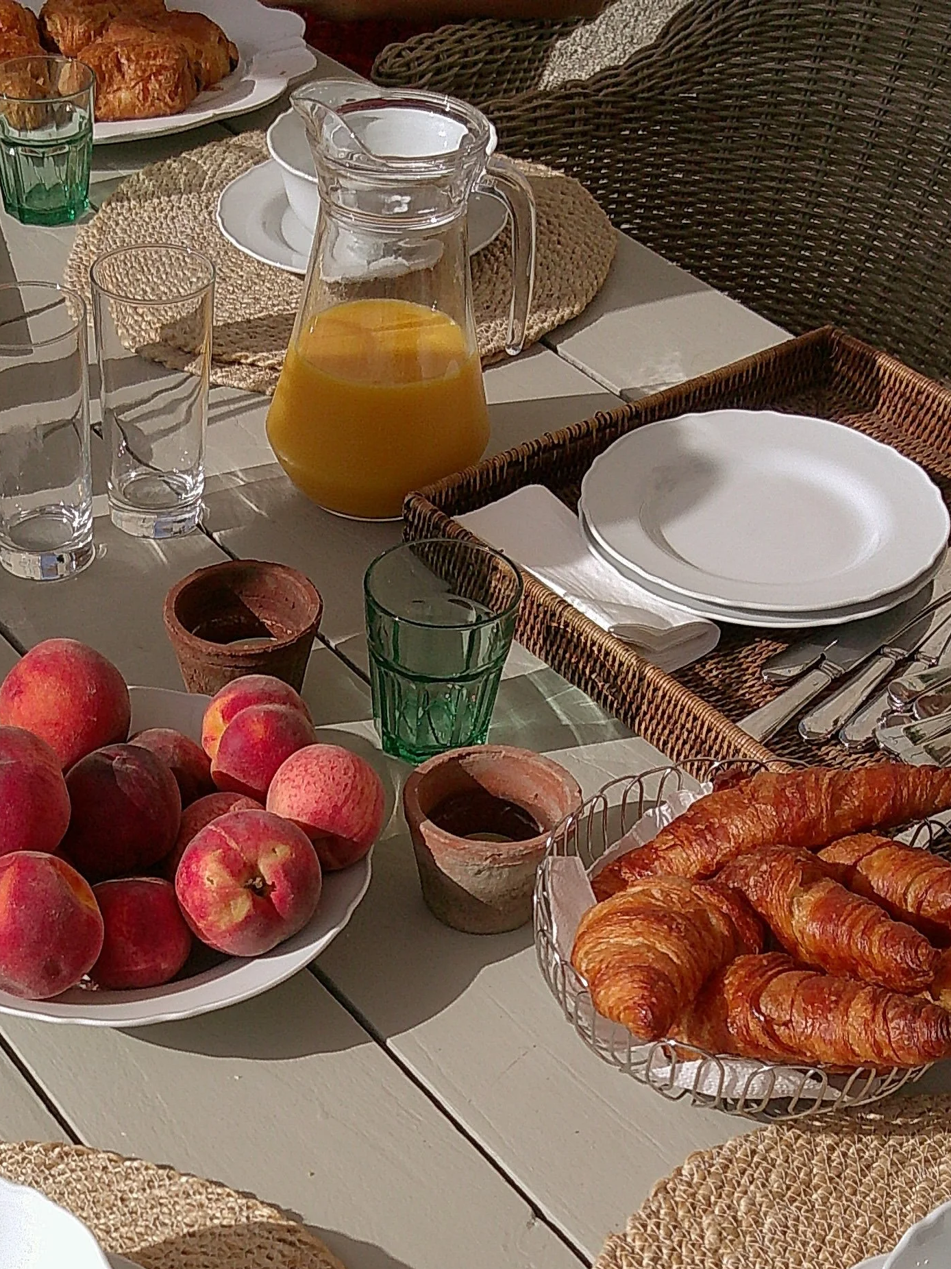 A breakfast table with a pitcher of orange juice, plates, glasses, apples, croissants, muffins, and croissants on a white table with woven placemats and chairs.