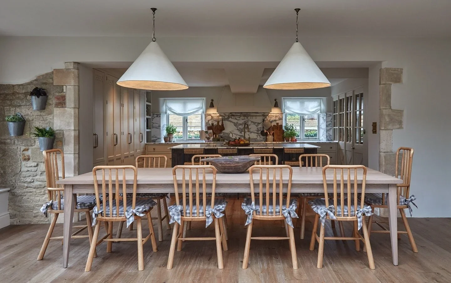 A spacious kitchen with a large wooden dining table and ten chairs with checkered bows, two hanging cone pendant lights, and a marble backsplash with open shelving and windows with curtains.