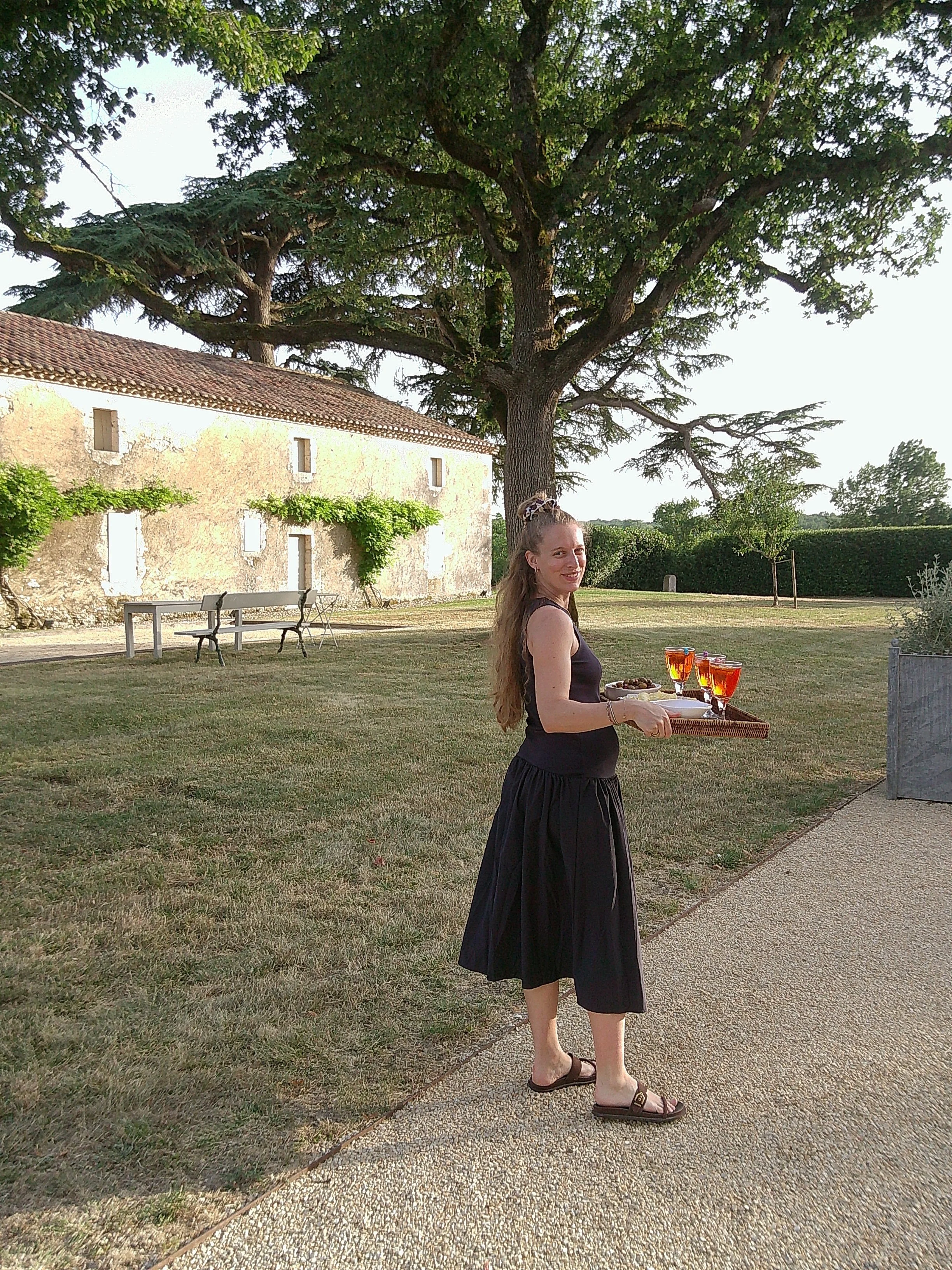 A woman with long, curly hair, wearing a black sleeveless top and a long black skirt, is holding a tray with glasses of orange beverages and a plate of snacks outdoors near a stone building with ivy and a large tree.