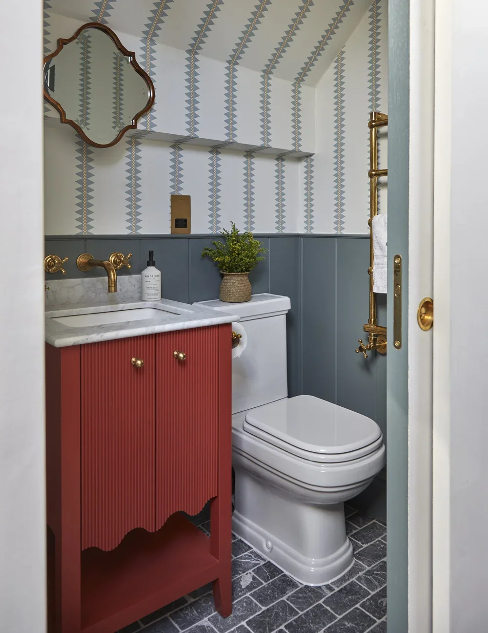 A small bathroom with patterned wallpaper, a white toilet, a red vanity with a marble top, a gold faucet, a potted plant, a mirror, and gold fixtures.