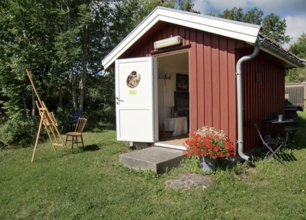 Et lite, rødt skur med åpen dør, omgitt av grønt gress og trær. Ute på venstre side står tre stoler, og på høyre side står et lite bord. Det er blomster i en stor potte foran skuret, og det er en nedløpsrør på siden av skuret.