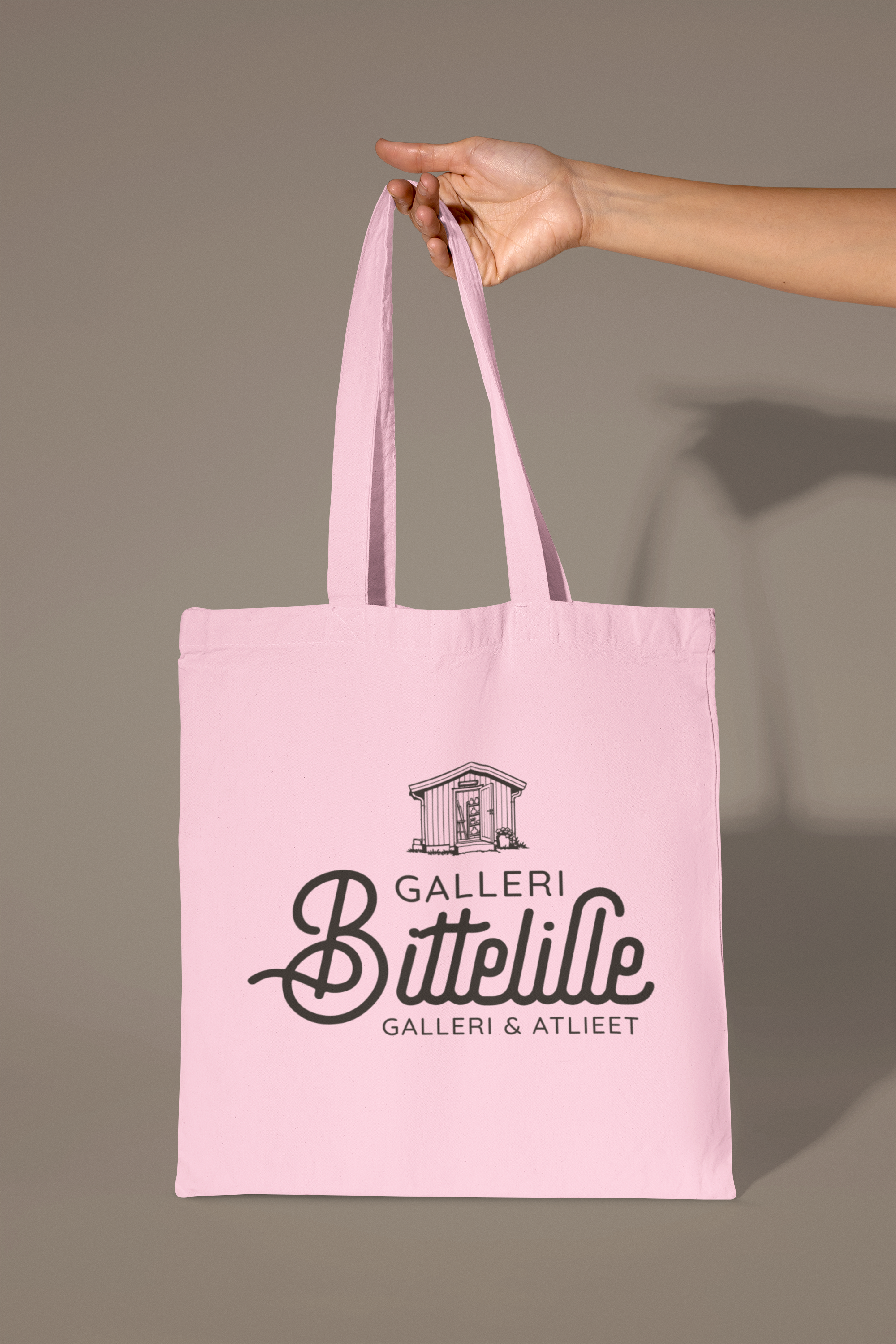 En person holder en rosa stoffveske med teksten "Galleri Bittelille" og en grafikk av en liten bygning, mot en grå bakgrunn.