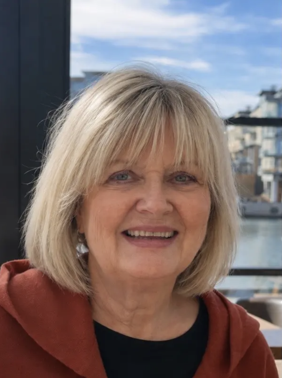 En fornøyd eldre kvinne med blondt hår og blå øyne, iført en rød genser, foran et vindu med en byhage i bakgrunnen.