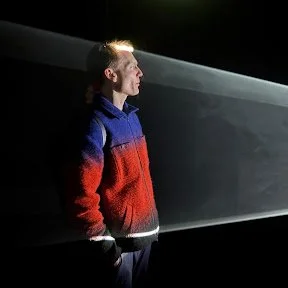 Young man with spiked hair and standing in profile in front of a beam of light.