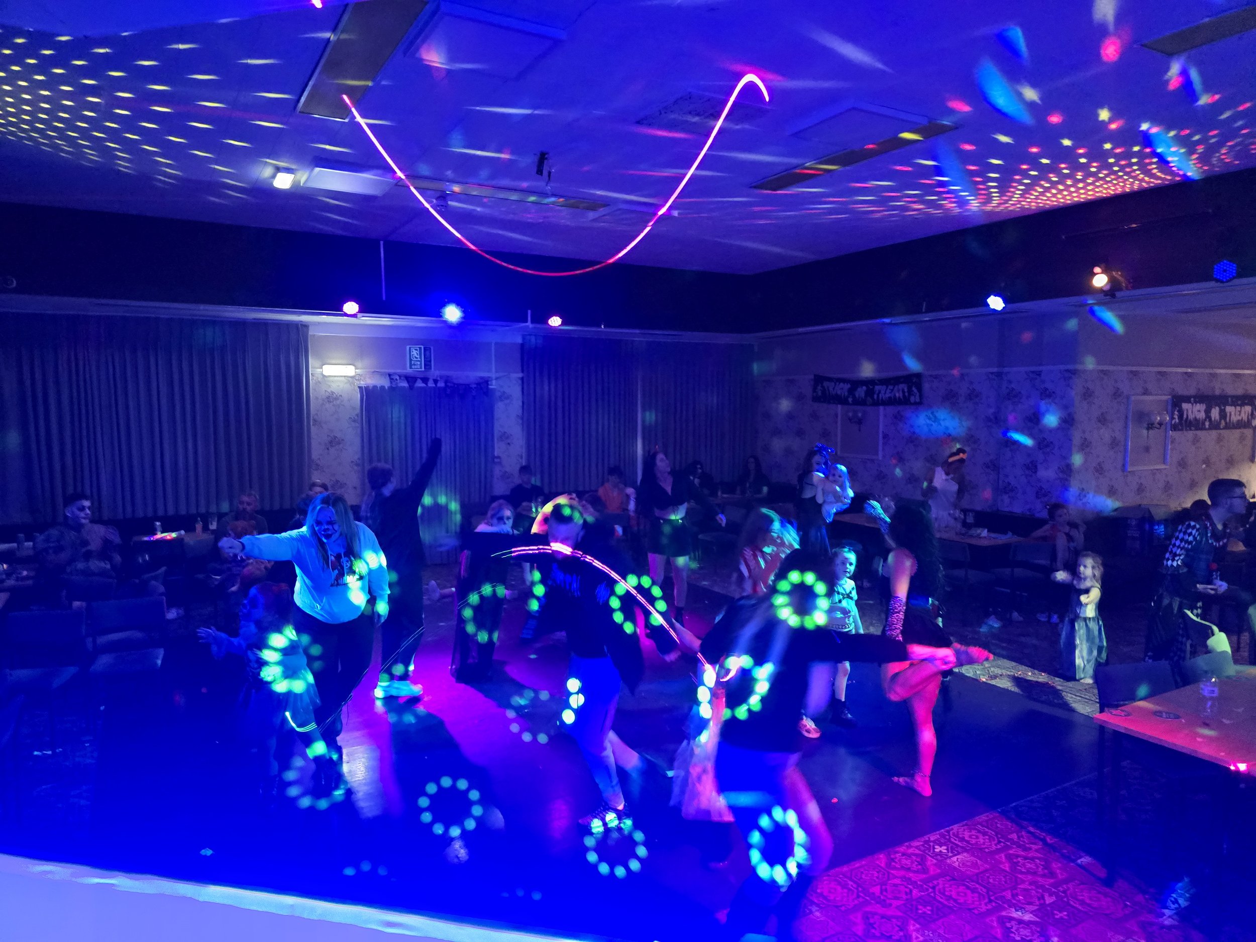 People dancing and playing with glow sticks at a party or event in a dimly lit room with colorful lights and laser patterns on the ceiling.