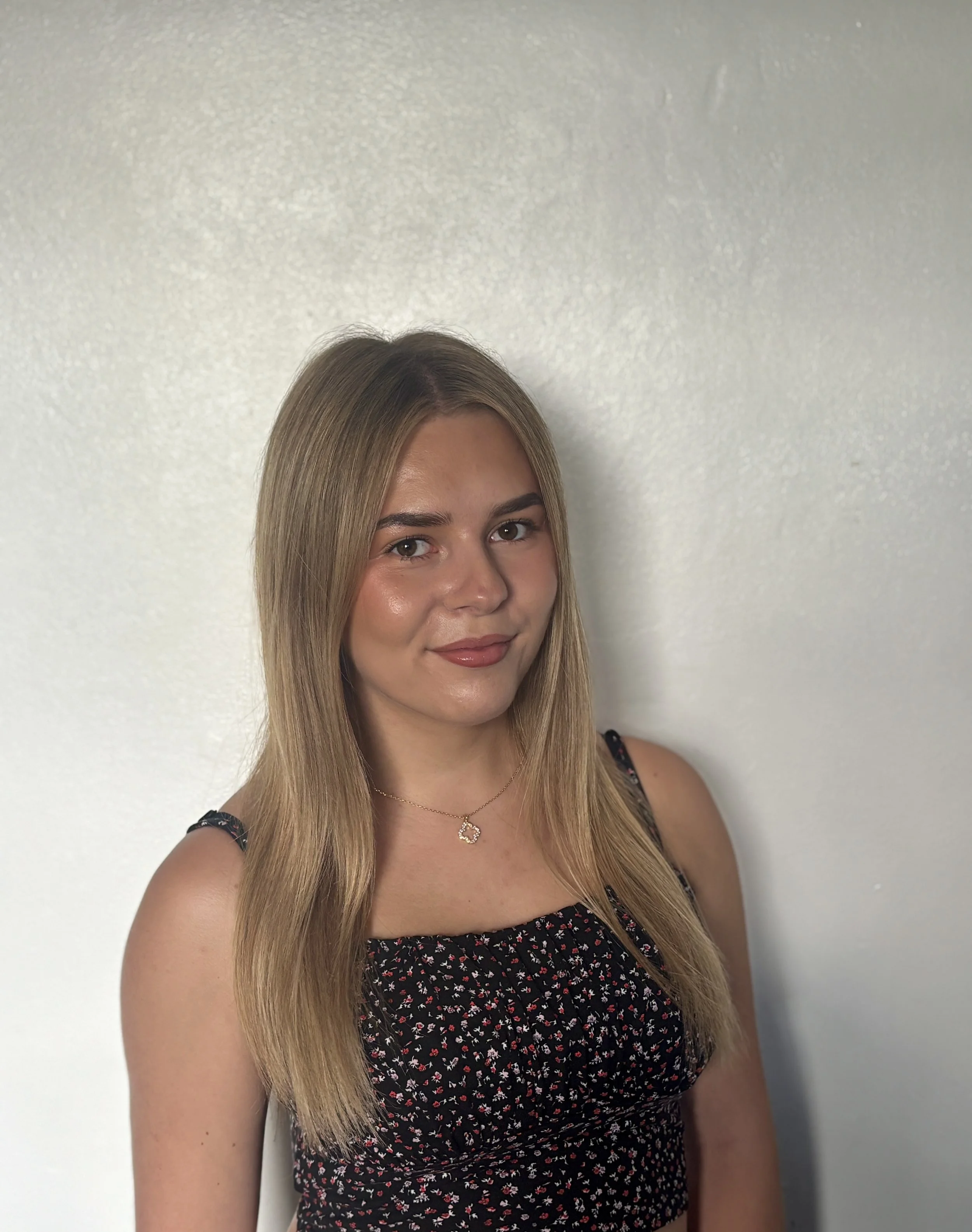 A young woman with long blonde hair wearing a black dress with a red and white floral pattern, a gold necklace with a heart-shaped pendant, standing against a plain white wall.