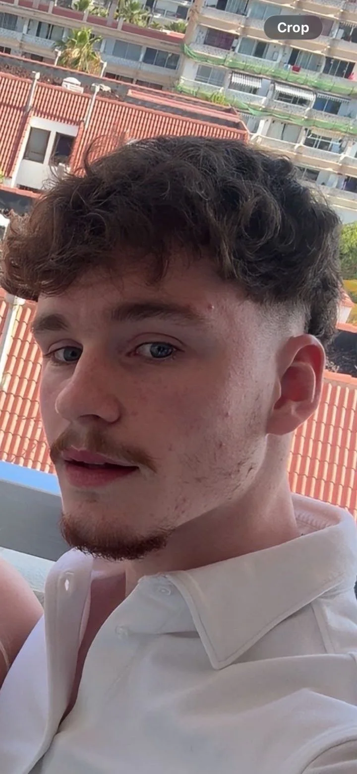 Close-up of a young man with curly hair, light stubble, blue eyes, and a white collared shirt, sitting on a balcony with a cityscape of apartment buildings and a rooftop with red tiles in the background.