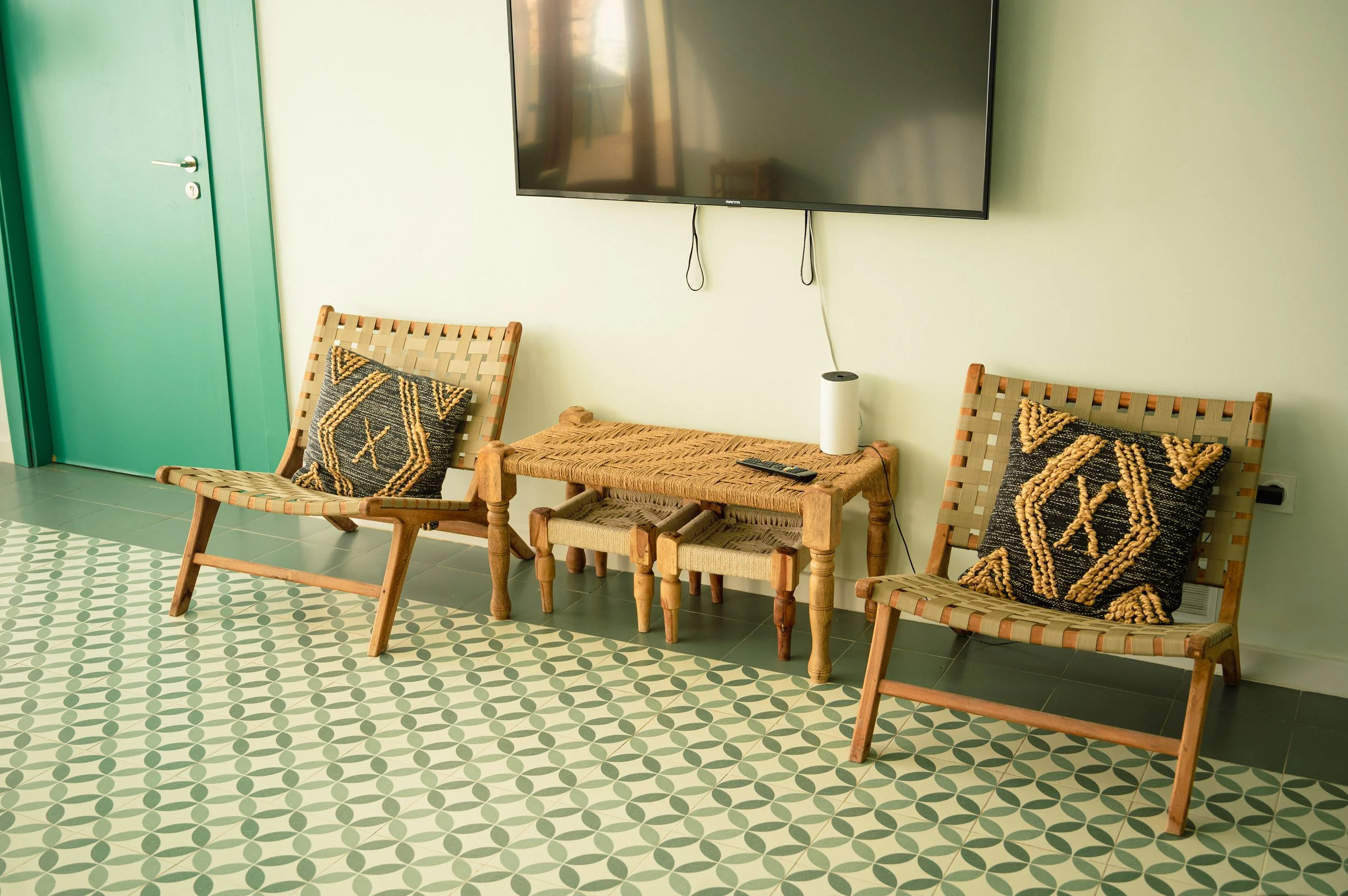 Sala con dos sillas de madera, cojines decorativos, mesa de madera, televisión de pantalla plana, ventilador inteligente y pared verde.