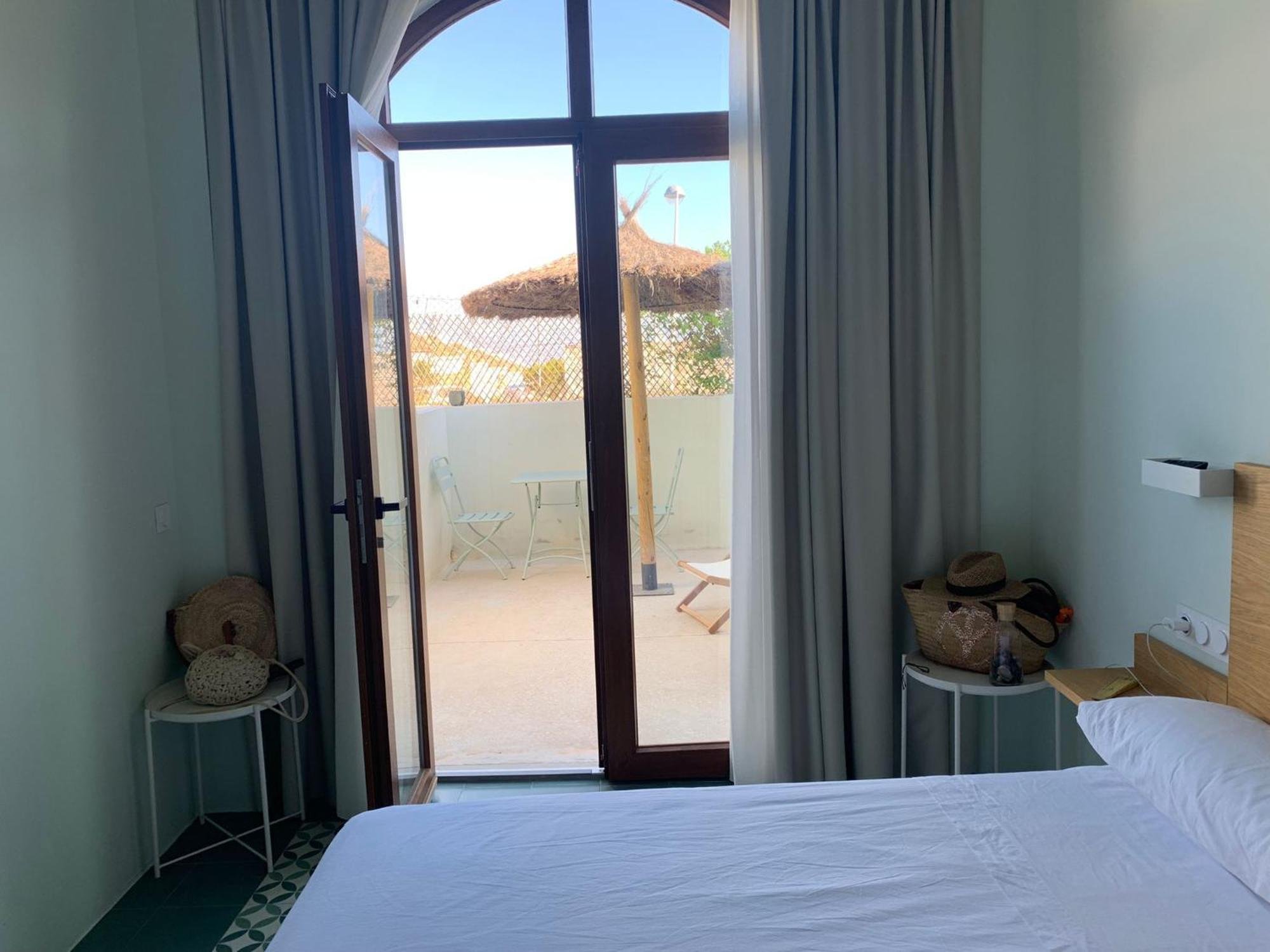 Vista de una cama en una habitación con paredes verdes, ropa de cama blanca, mesas auxiliares con sombreros de playa, y una puerta de cristal que lleva a un balcón con sillas y sombrillas de paja, y vista del cielo y árboles.