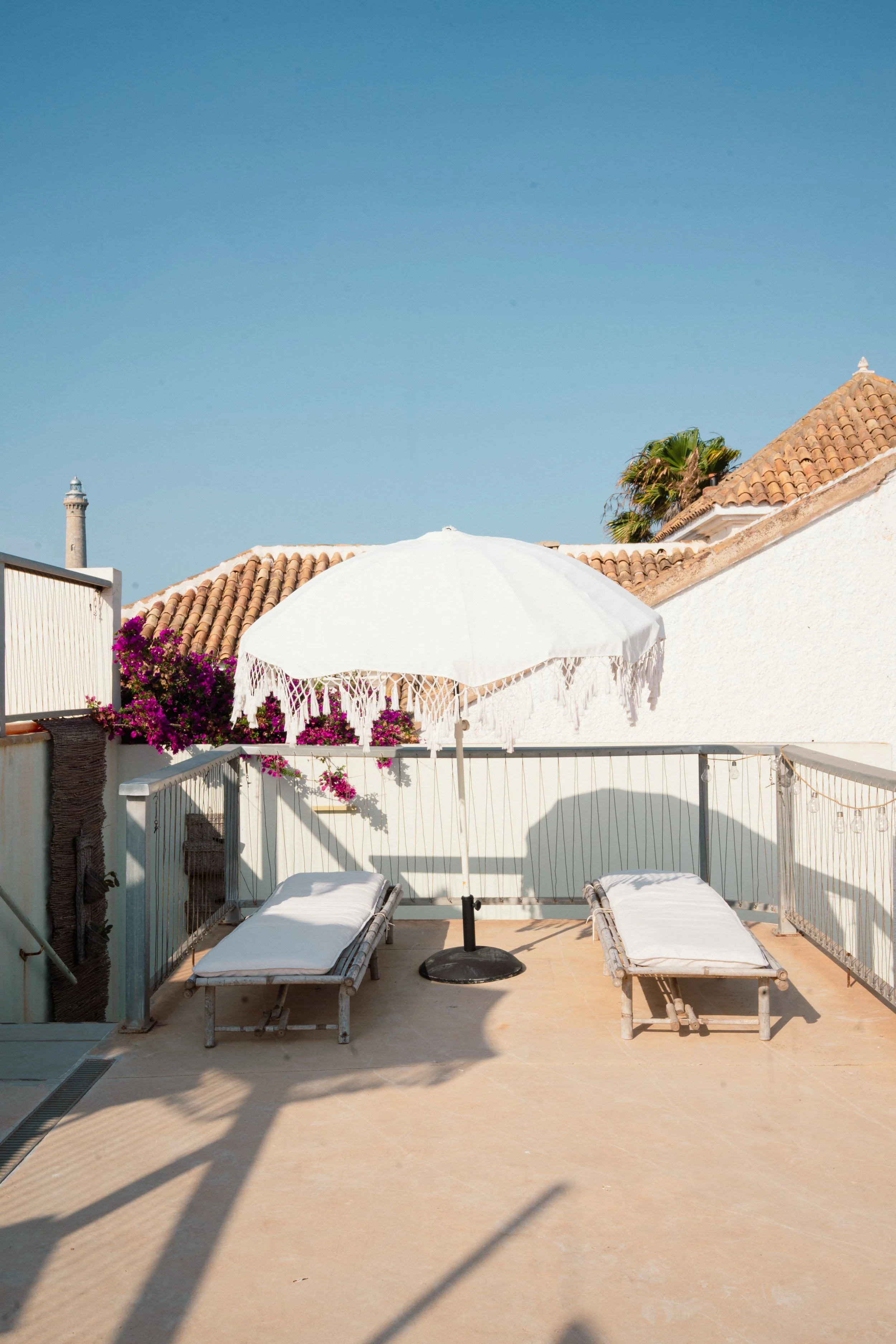 Una terraza con dos camastros, una sombrilla blanca con bordes de encaje, una cerca blanca con flores fucsias, un techo de tejas y un cielo azul despejado.