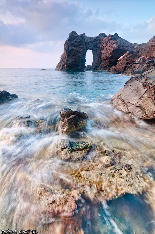 Formación rocosa en el mar con un arco natural en la estructura, olas en primer plano y cielo nublado.