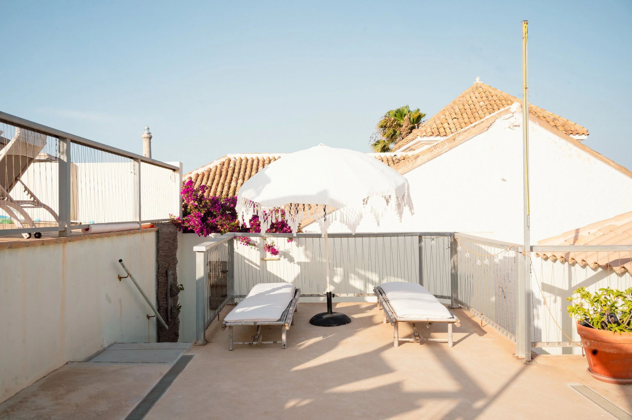 Terraza de azotea con sillas y sombrilla blanca, paredes blancas, cerámica en el suelo, plantas en macetas, y techos de tejas en casas cercanas, con cielo despejado.
