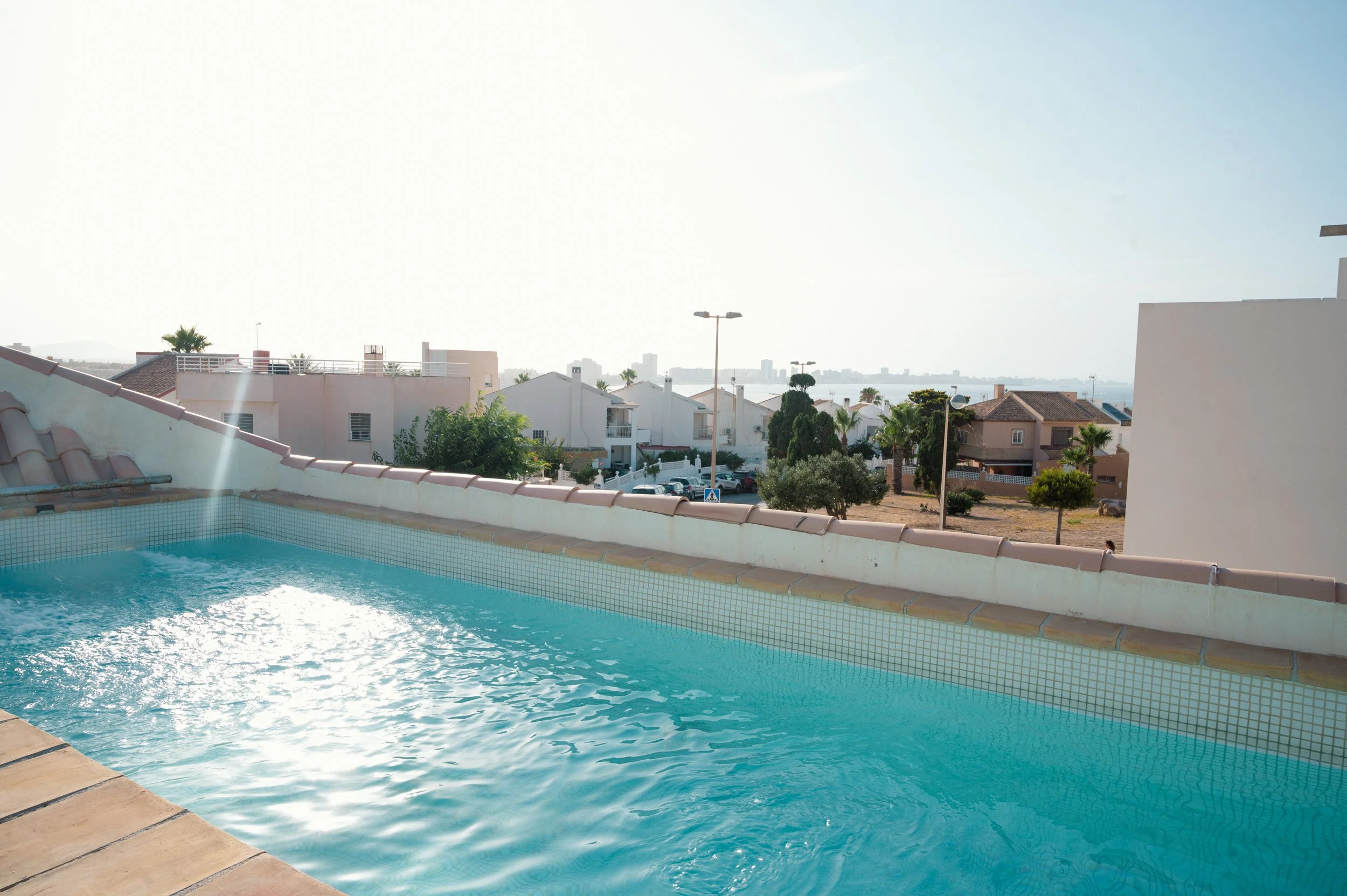 Piscina con vista a casas y edificios, con árboles y farolas, en un día soleado y despejado.