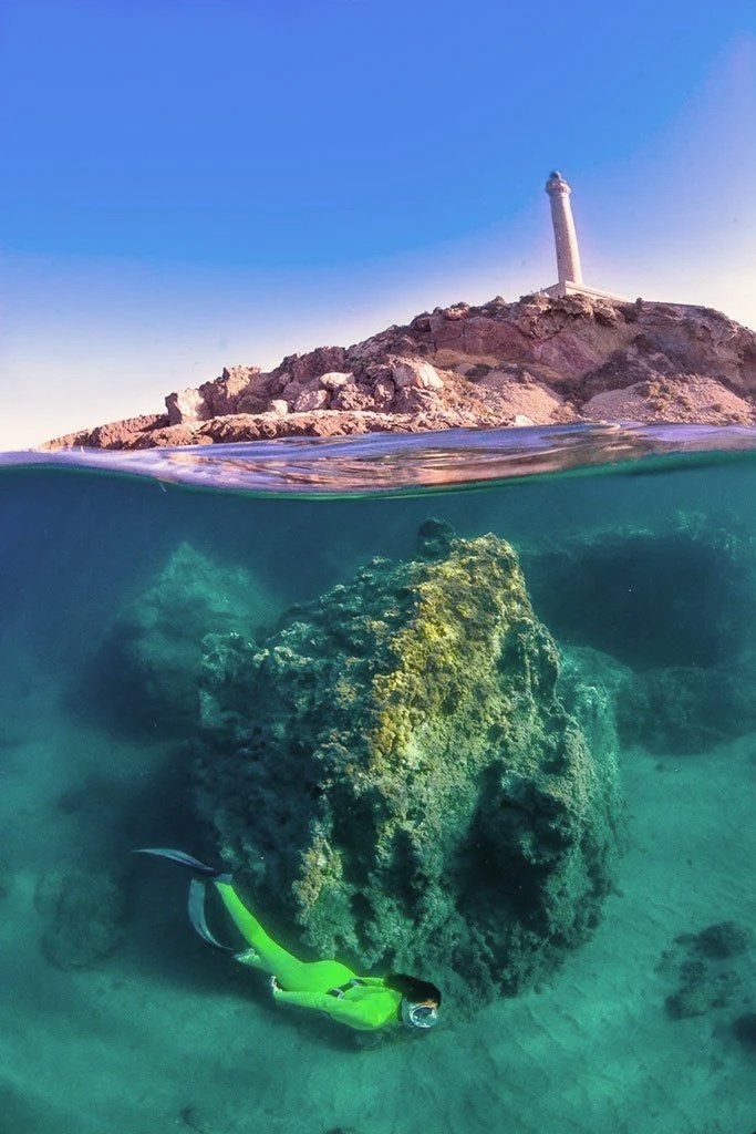 Vista dividida entre tierra y mar. En la parte superior, un faro en un promontorio rocoso bajo un cielo azul. En la parte debajo del agua, una roca con coral y un buzo con traje y máscara explorando el fondo marino.