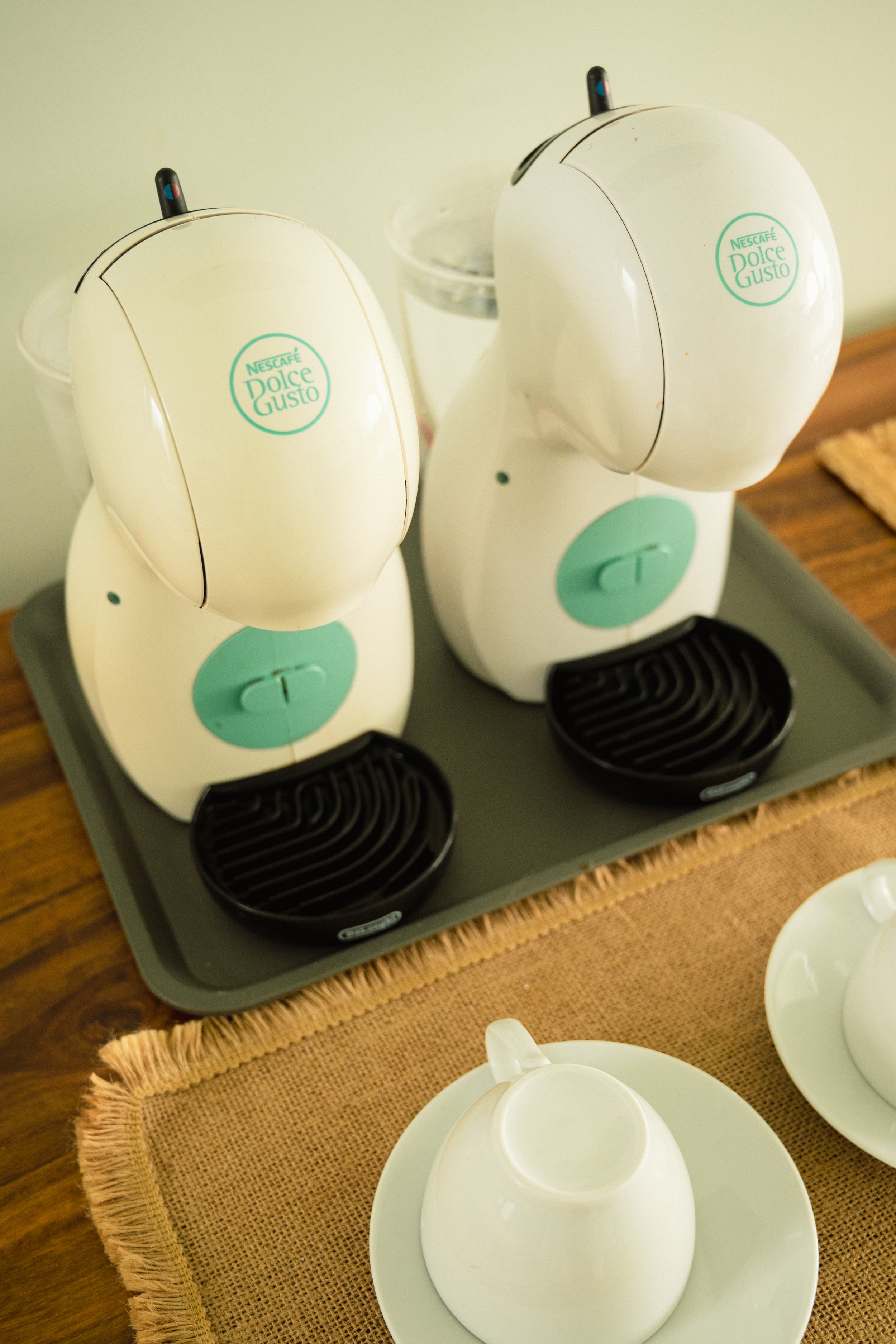 Dos cafeteras blancas con logo de Nescafé Dolce Gusto, sobre un plato gris en una mesa de madera con mantel de yute, acompañadas de tazas blancas.