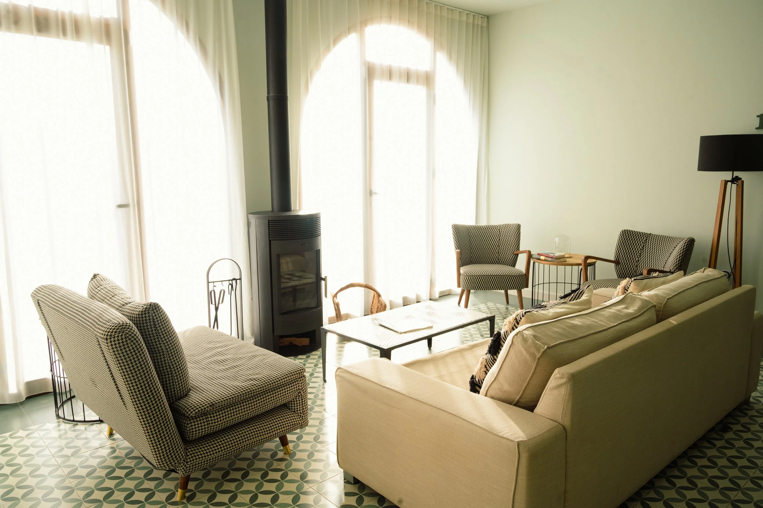 Sala de estar con sillones de tela a cuadros, una mesa de centro, una estufa de leña, cortinas blancas y piso de baldosas con patrón geométrico.