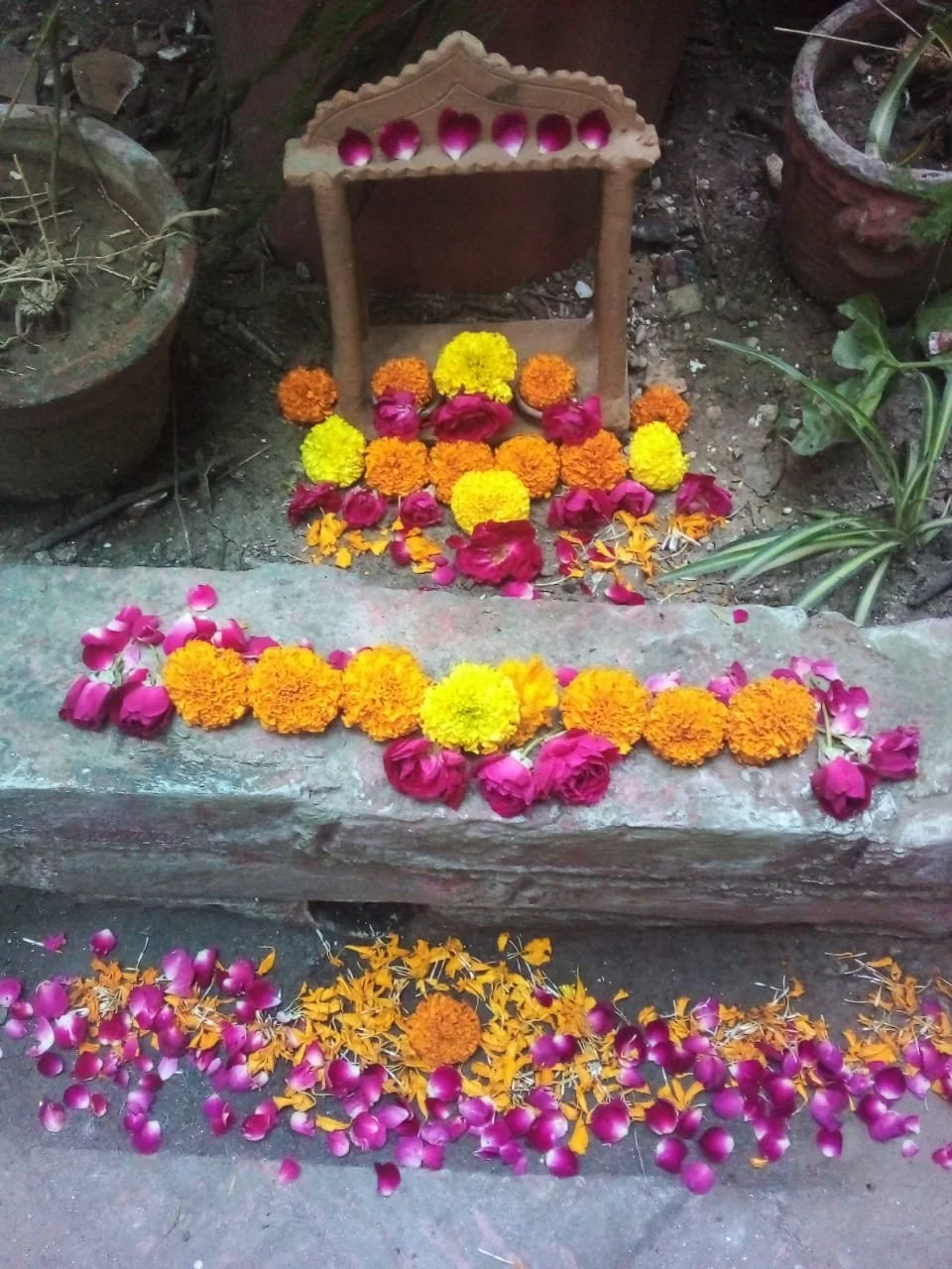 A small outdoor shrine with colorful flower arrangements including marigolds, roses, and petals, placed on the steps and surrounding area, with potted plants nearby.