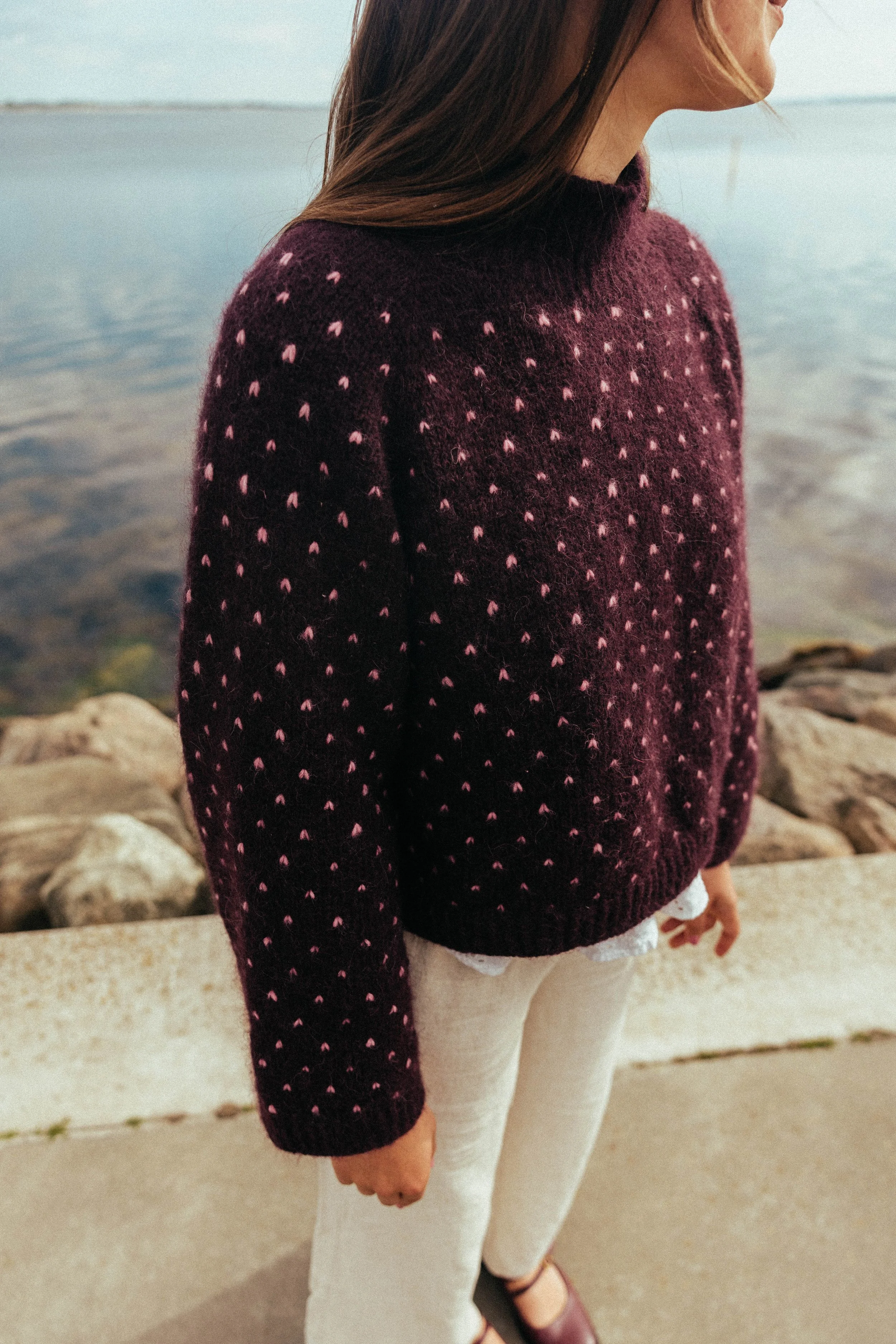 En person står ved en kyst med store sten og havet i baggrunden. Personen har på en mørkerød sweater med lyserøde prikker, hvide bukser og sandaler.