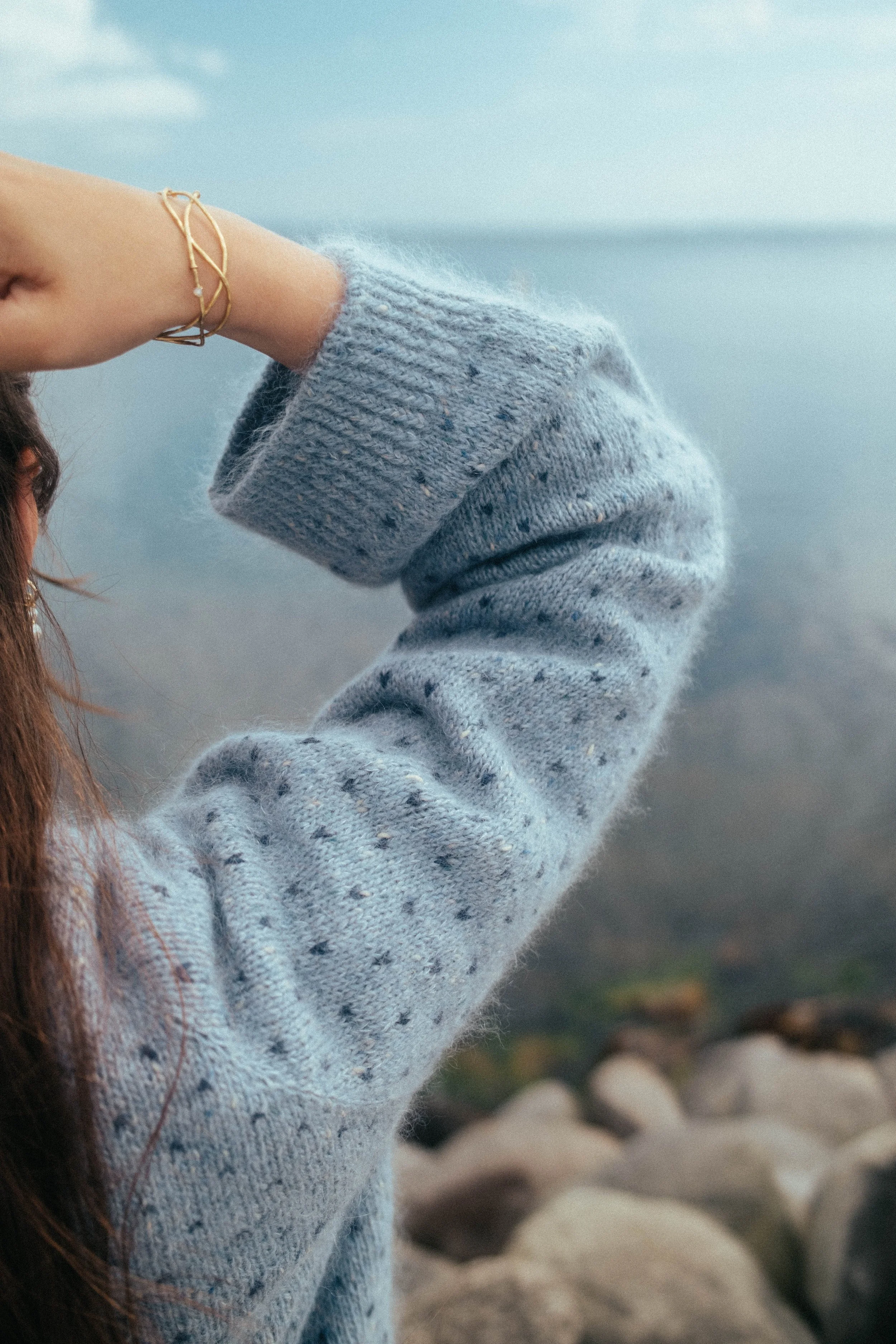 Nær billede af en persons arm som er bøje, iført en blå strikket sweater, med en gullarmbånd. Flot vand i baggrunden