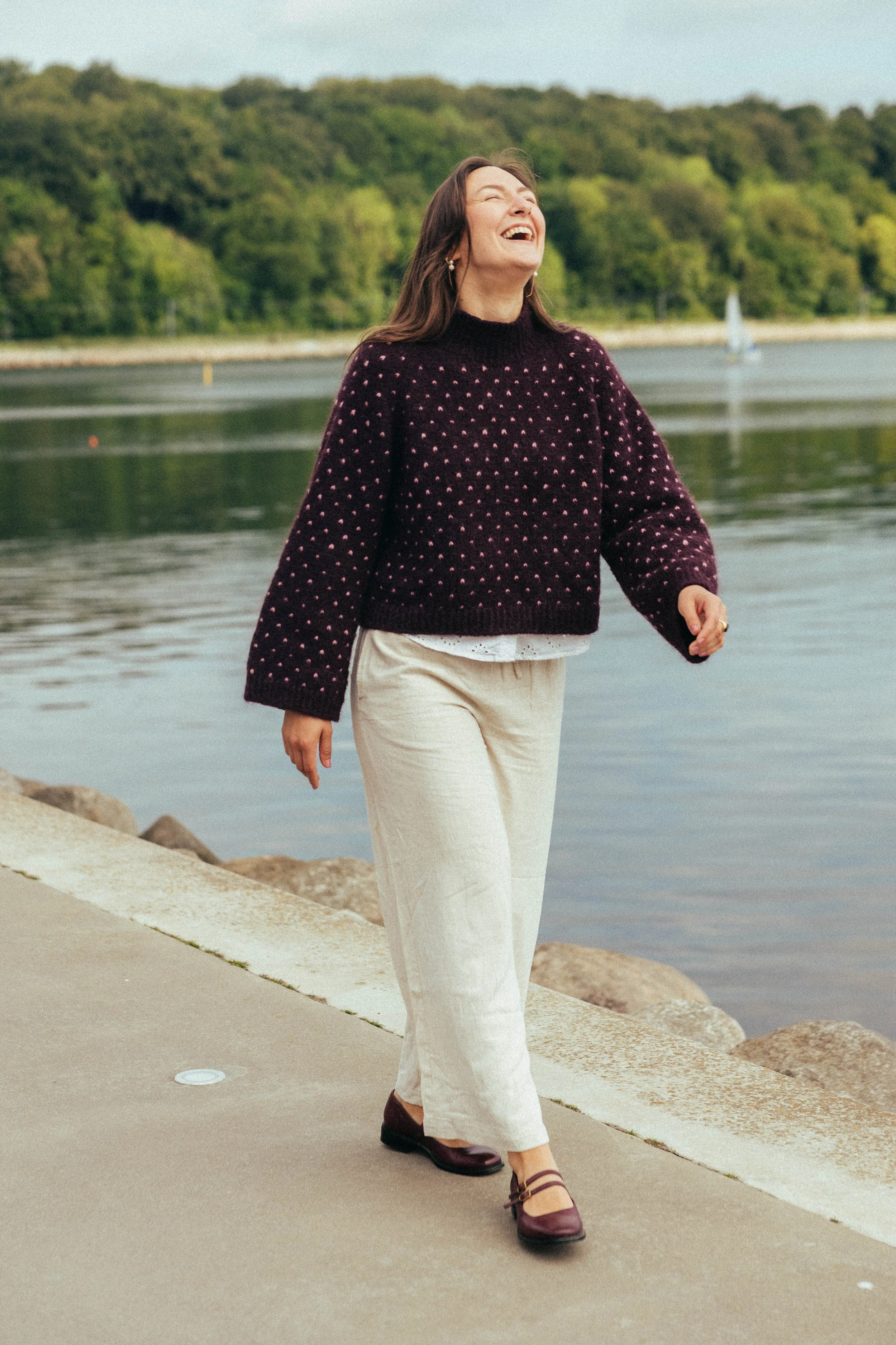 En kvinde i en plettet sweater, hvide bukser og vinrøde sko går ved en sø med grøn skov i baggrunden.