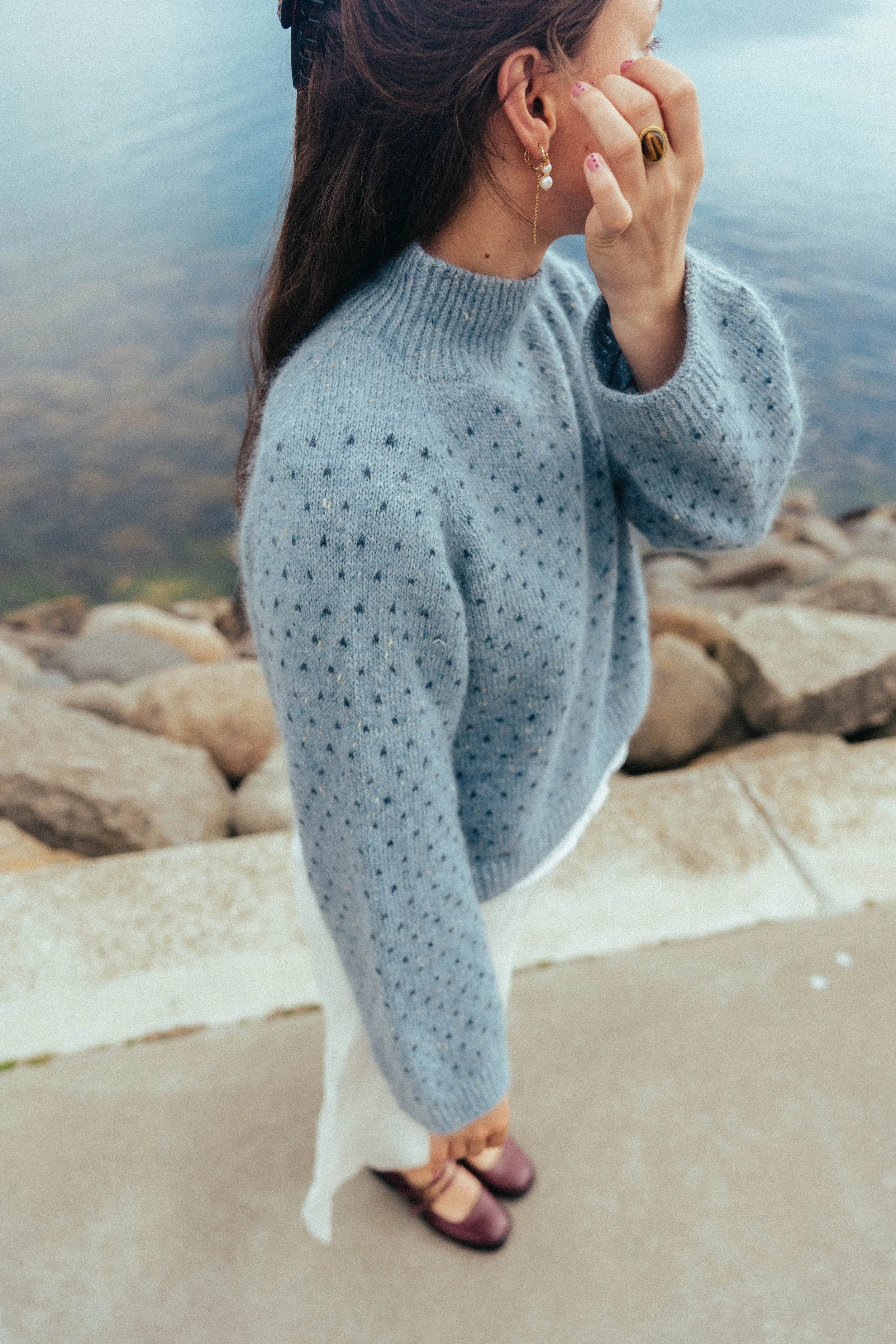 En kvinde i en blå sweater står ved vandet, med ryggen mod kamera. Hun har langt, mørkt hår og bærer øreringe og ringe. Hun har sandaler og står på en sten- og betonflise belægning.