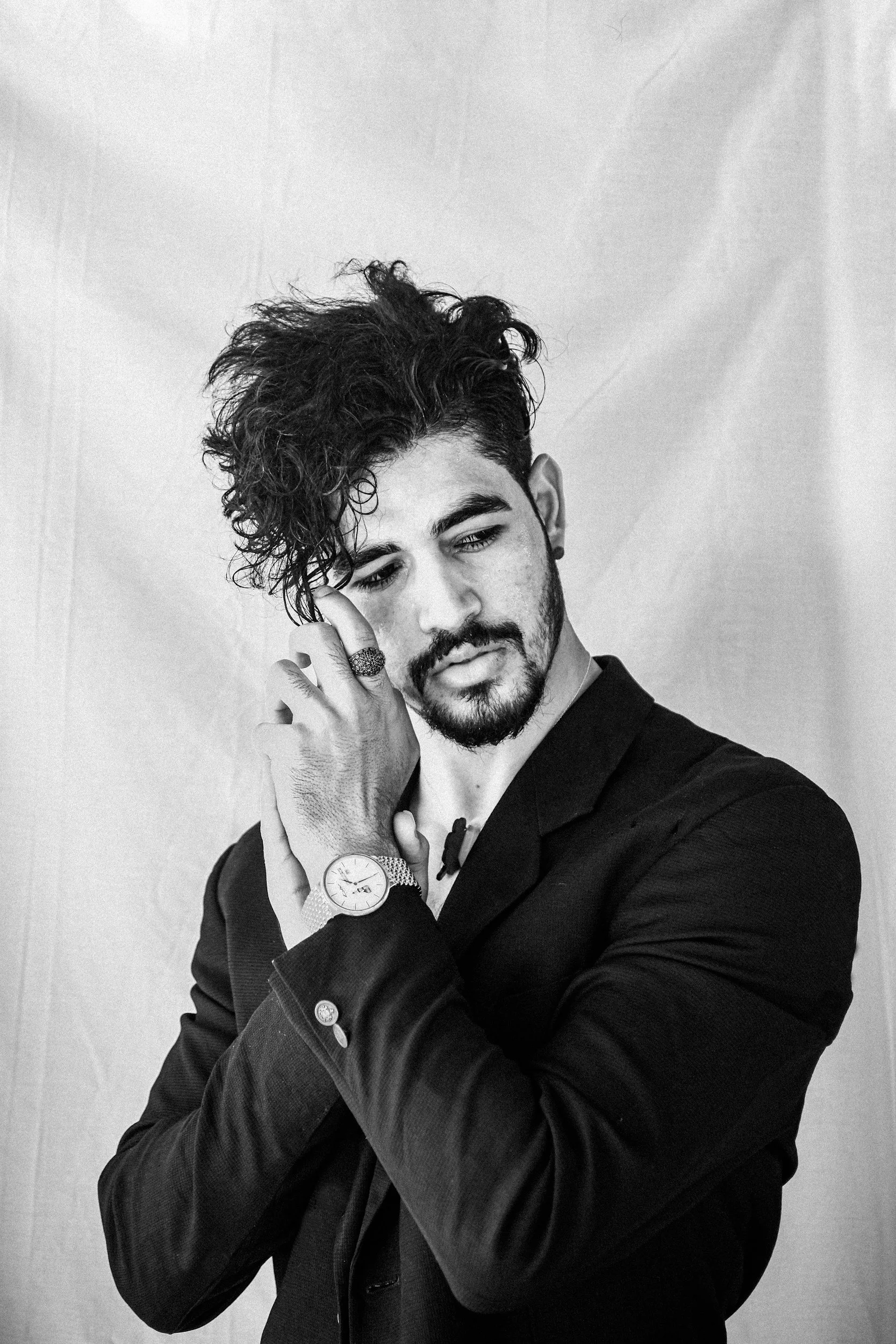Black and white portrait of a young man with curly hair, beard, and mustache, wearing a dark suit jacket, a watch, and rings, touching his face thoughtfully against a plain background.