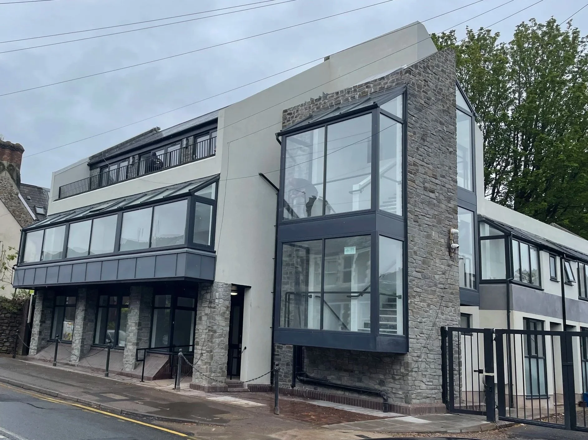 Modern multi-story building with large glass windows, stone and white exterior walls, located on a street with a designated sidewalk and a black metal fence.