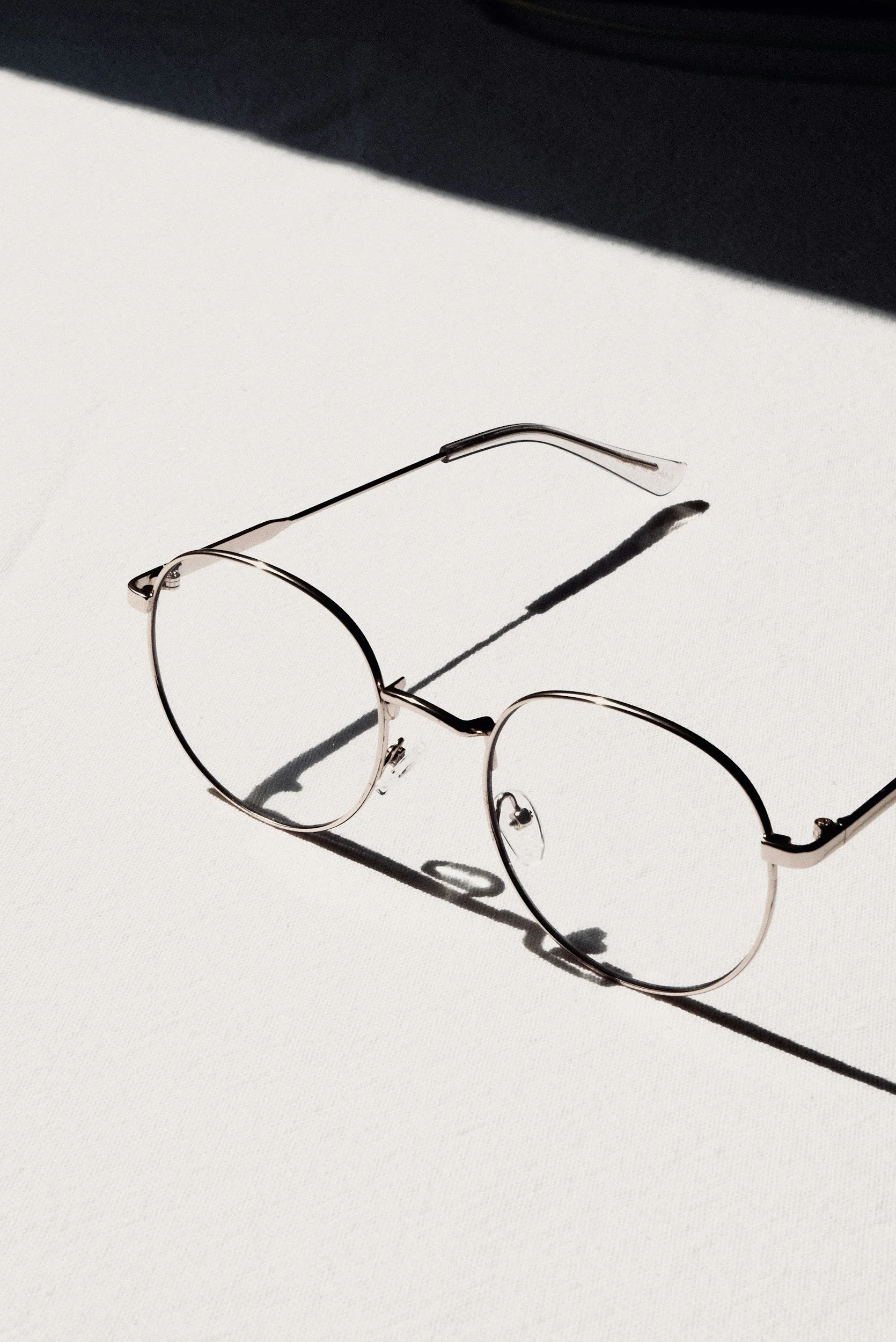 A pair of round eyeglasses with thin metal frames resting on a white surface, casting a shadow.