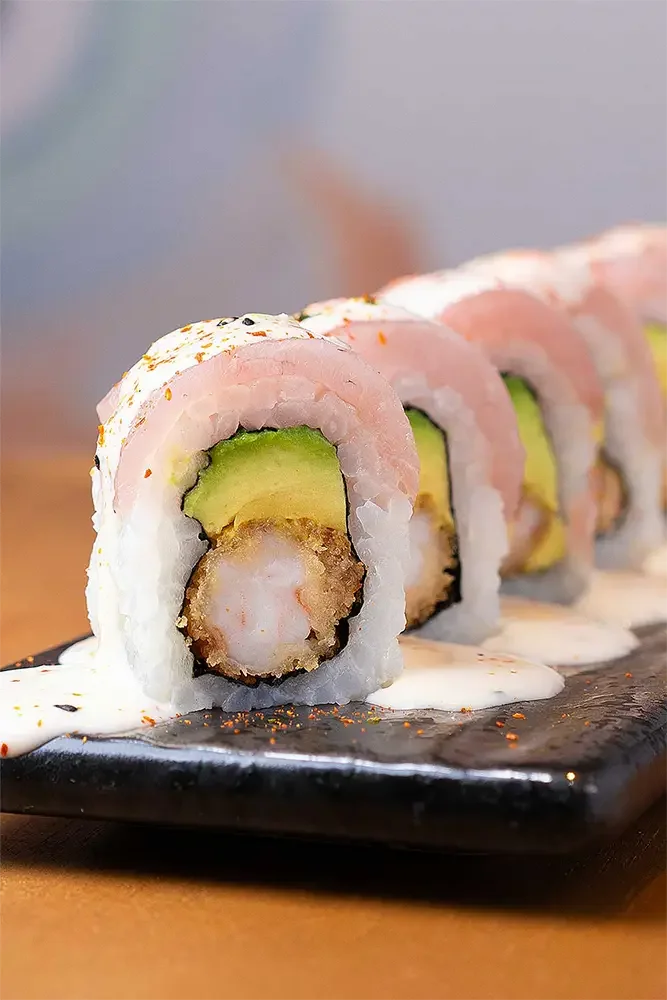 Rollo de sushi con camarón frito, aguacate y cecina en un plato negro, con salsa blanca y especias por encima.