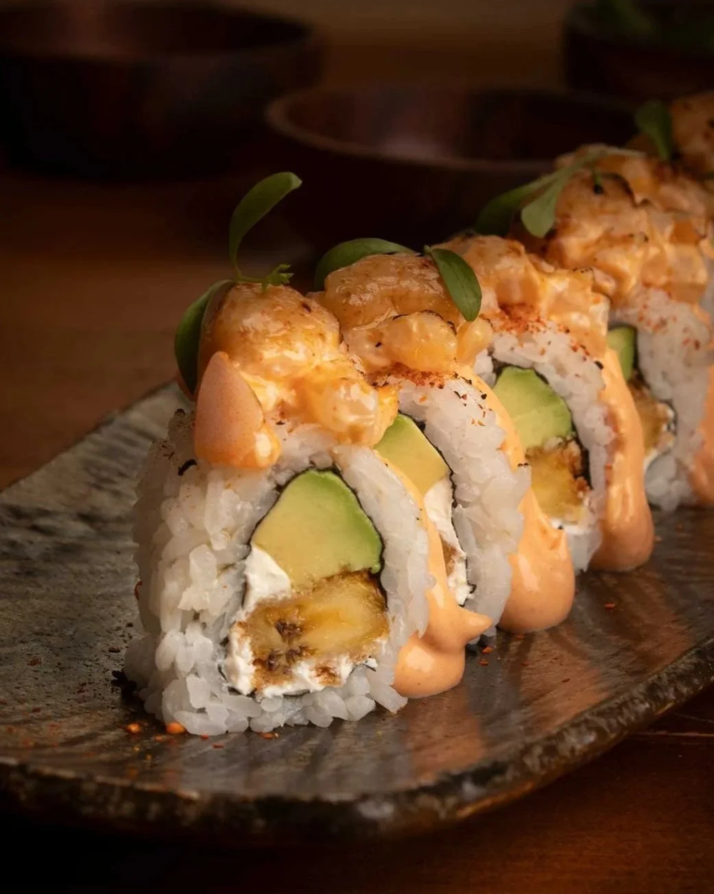 Rollos de sushi con aguacate, crema y salsa picante, decorados con hojas verdes sobre un plato de cerámica.