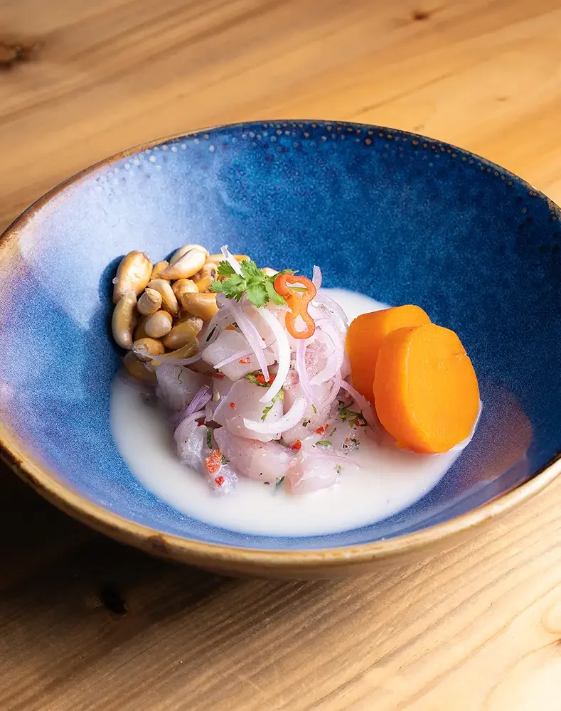 Pescado crudo en salsa de leche, con rebanadas de zanahoria, cebolla y cilantro, acompañado de maní y una rodaja de naranja en un plato azul